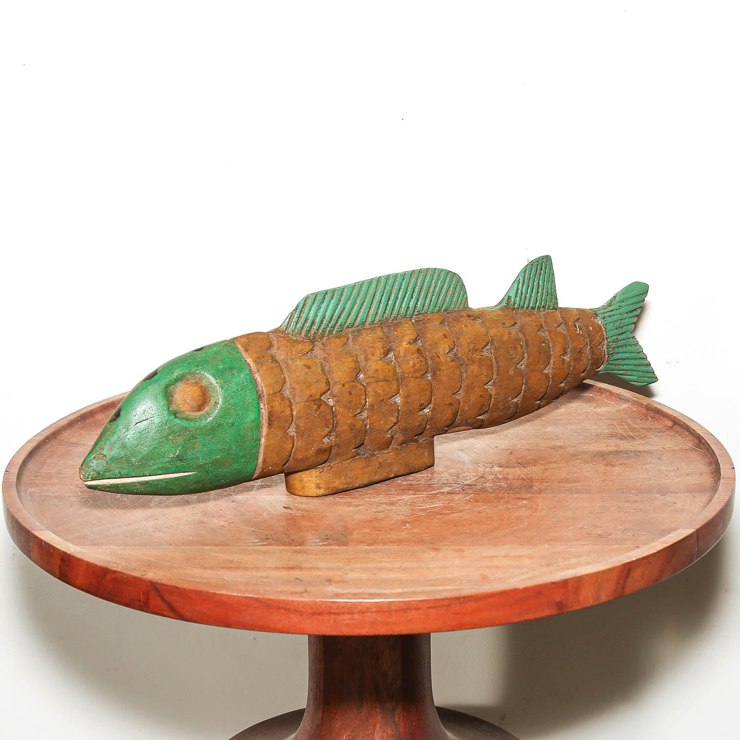 A hand-carved wooden fish figurine with a green head and fins, resting on a round wooden pedestal against a plain white background.