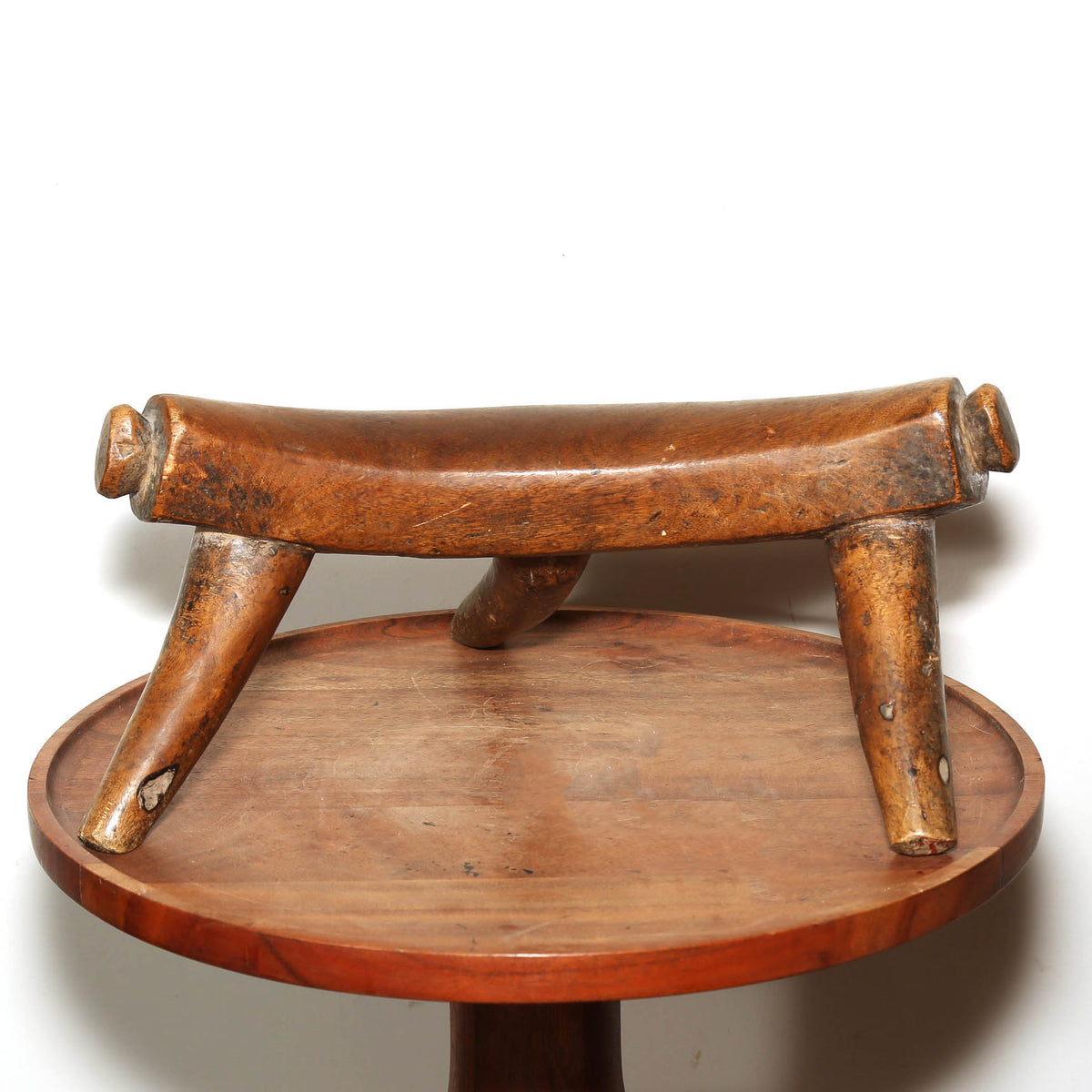 A rustic carved wooden stool with a curved seat and four legs, shown on a round wooden table against a plain white background.