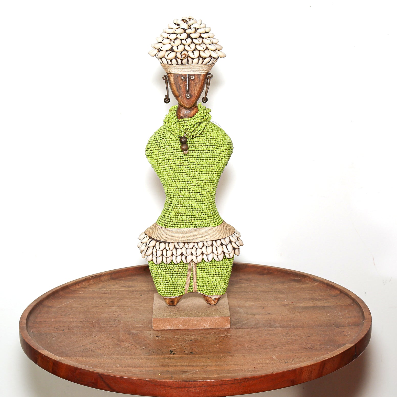 A handmade African-style beaded female figurine with a green textured dress, white shell skirt, and a shell-covered headdress, displayed on a round wooden tray against a white background.