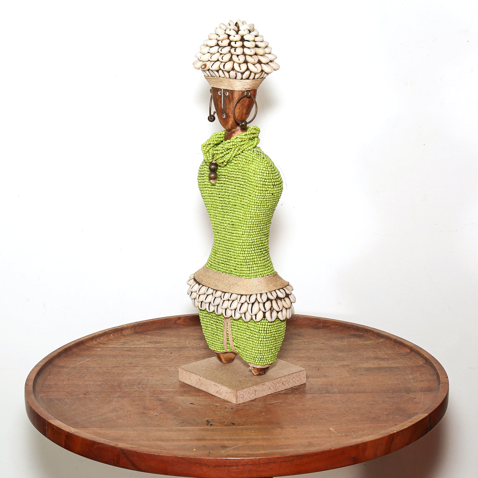 A handmade African-style wooden figurine of a woman wearing a bright green beaded dress and a skirt made of cowrie shells, displayed on a round wooden table against a white background.