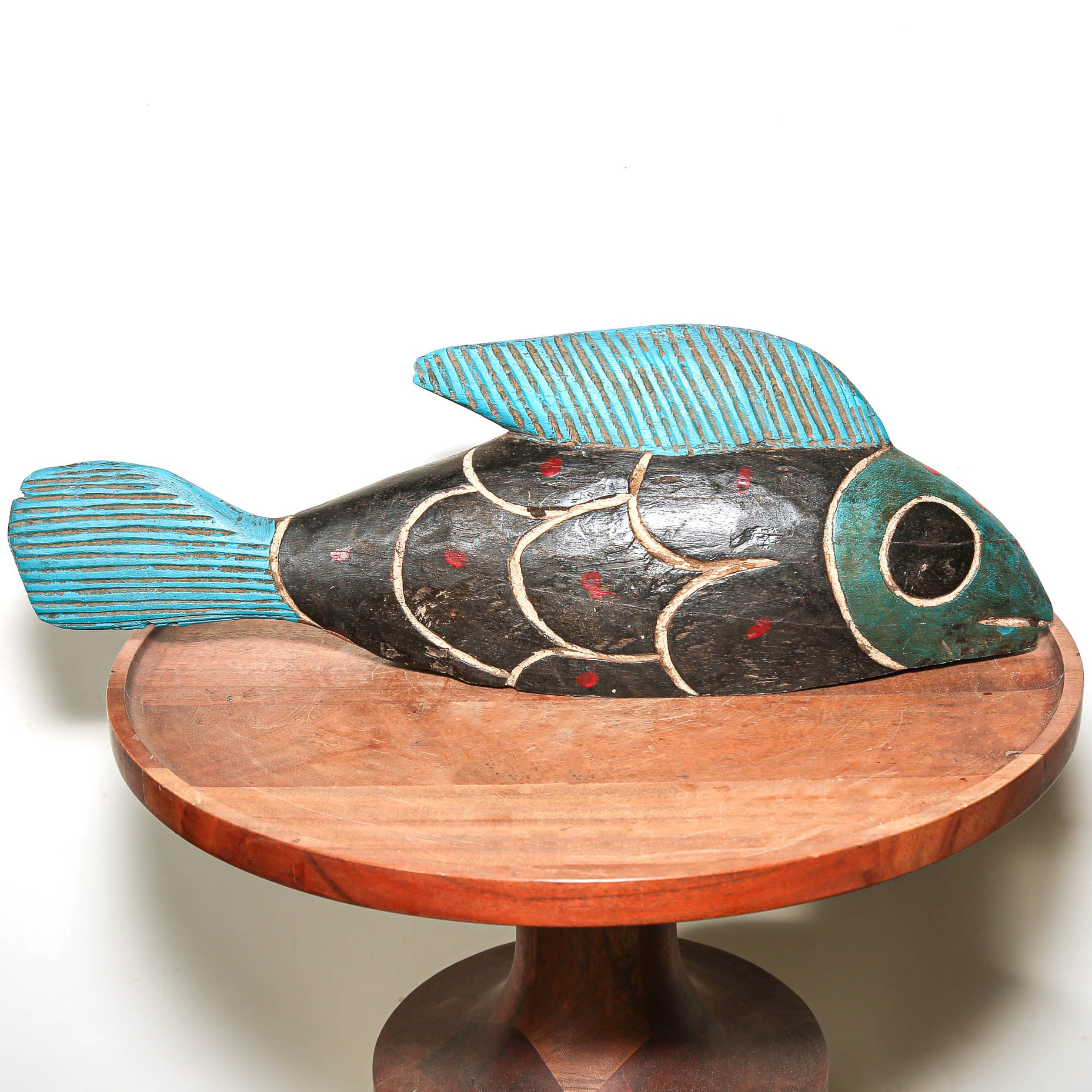 A hand-carved wooden fish sculpture with turquoise blue painted fins, a black body, and red and white painted details, displayed on a round wooden pedestal against a white background.