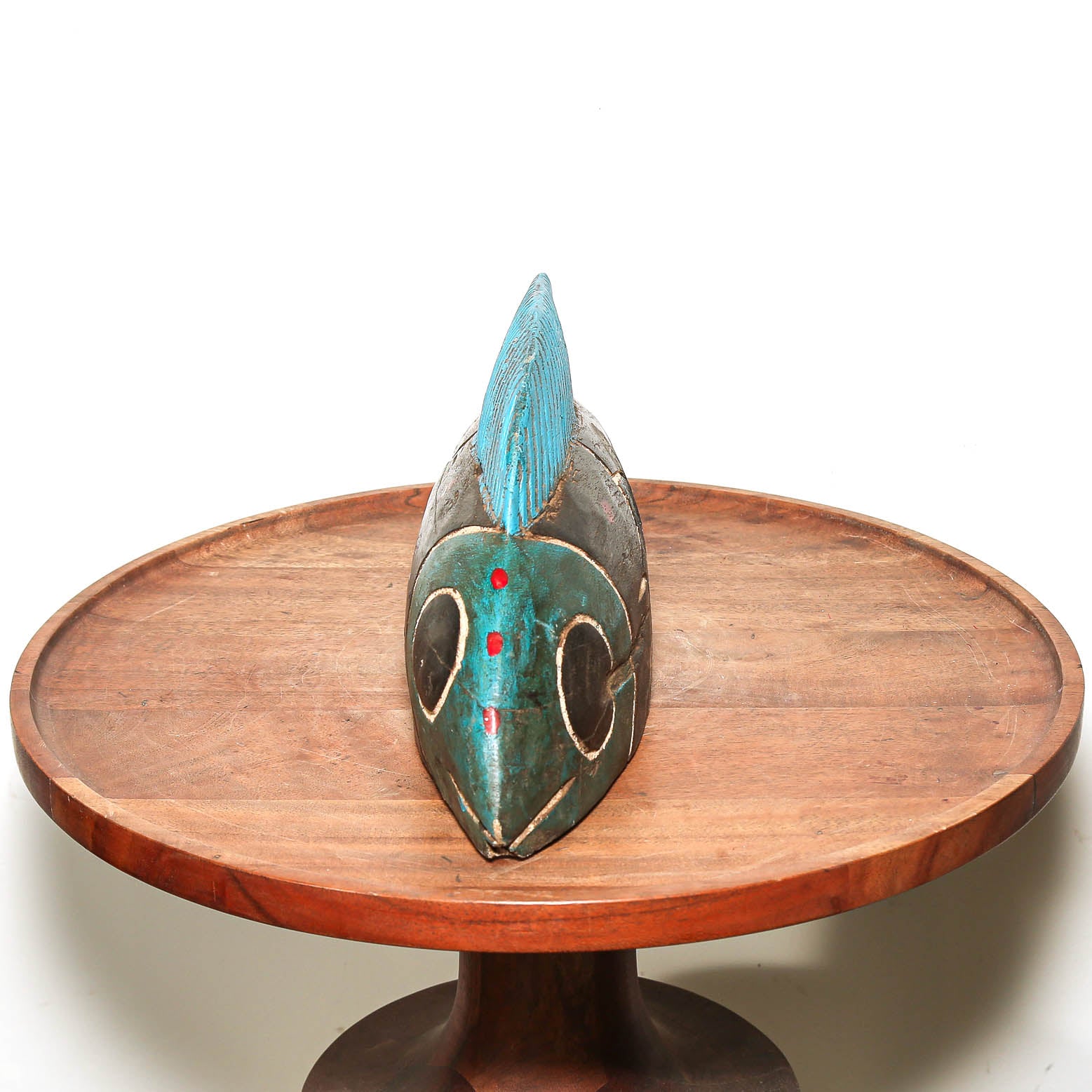 A carved African wooden mask with turquoise blue, green, and red accents, displayed on a round wooden pedestal against a white background.