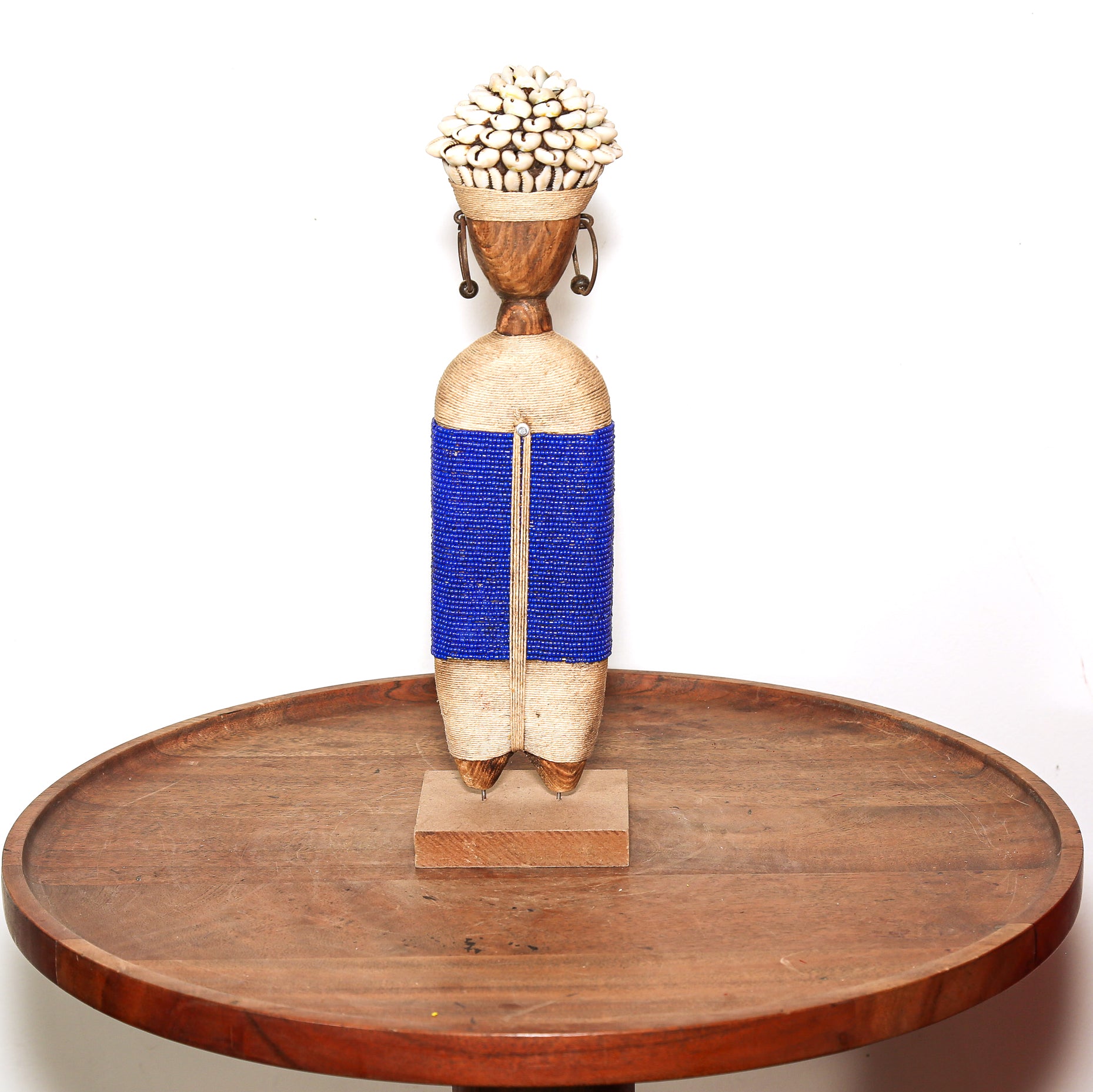 A handcrafted African-style wooden figurine with a blue beaded waist and a basket of small white shells on its head, displayed on a round wooden tray against a white background.