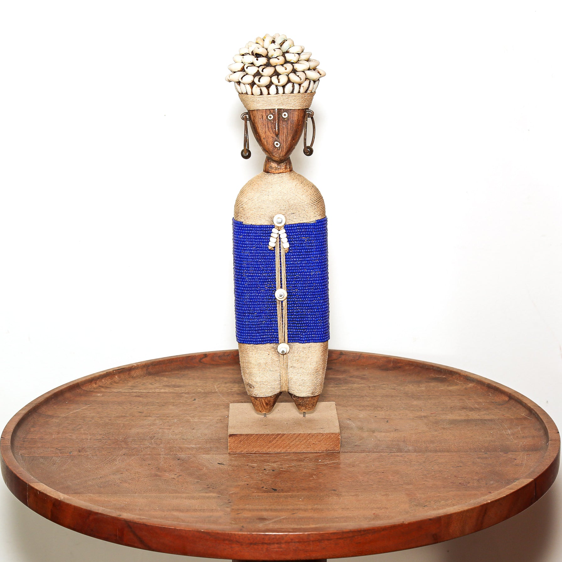A handcrafted African-style wooden figurine with a carved face, shell hair, beaded blue waist, and small wooden shoes, displayed on a round wooden tray against a white background.