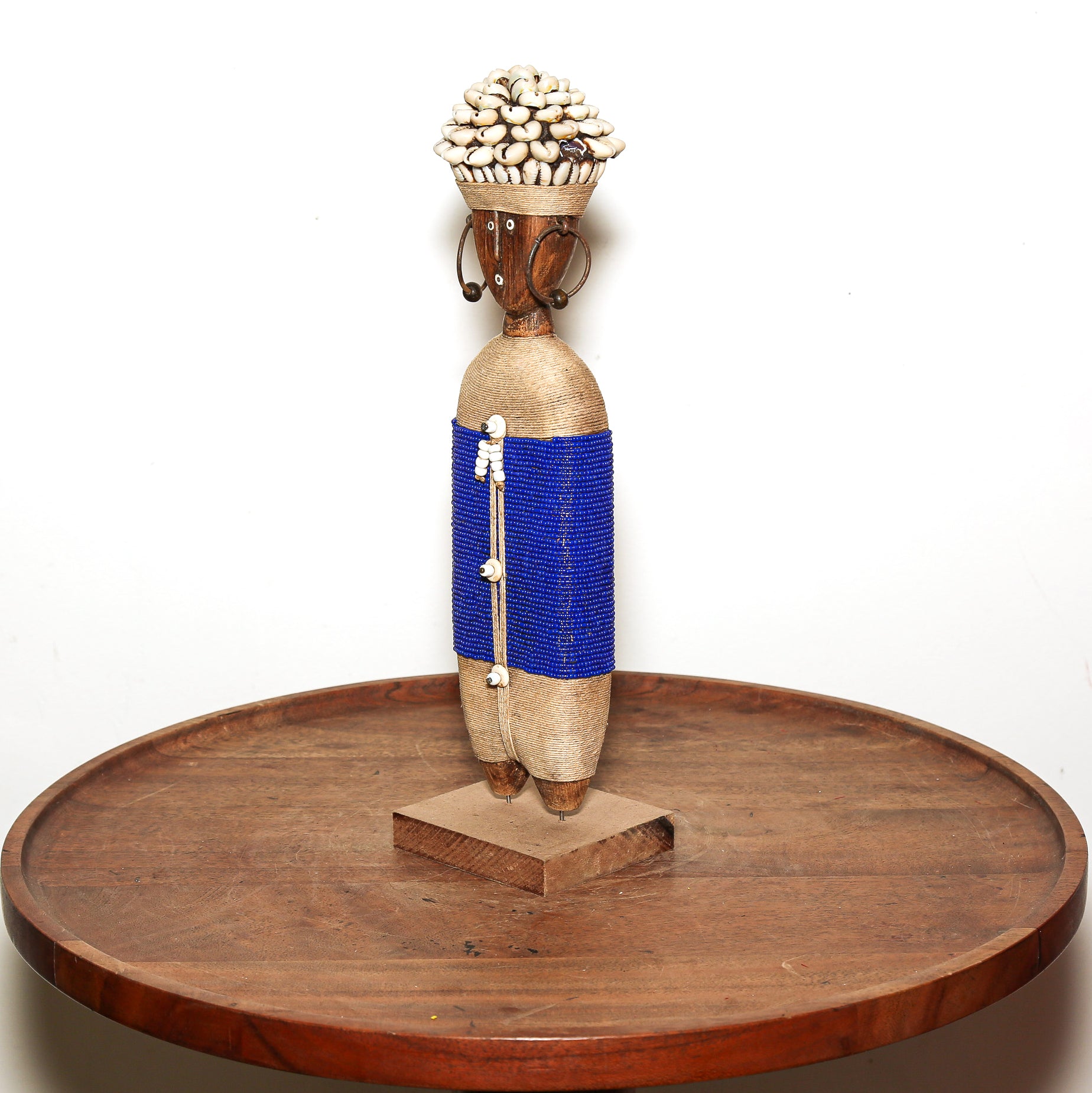 A handmade African-style decorative figurine on a round wooden table against a white background. The figurine features a natural jute body wrapped with a vibrant blue beaded waist band and a white beaded hanging ornament, topped with a woven basket holding white cowrie shells.