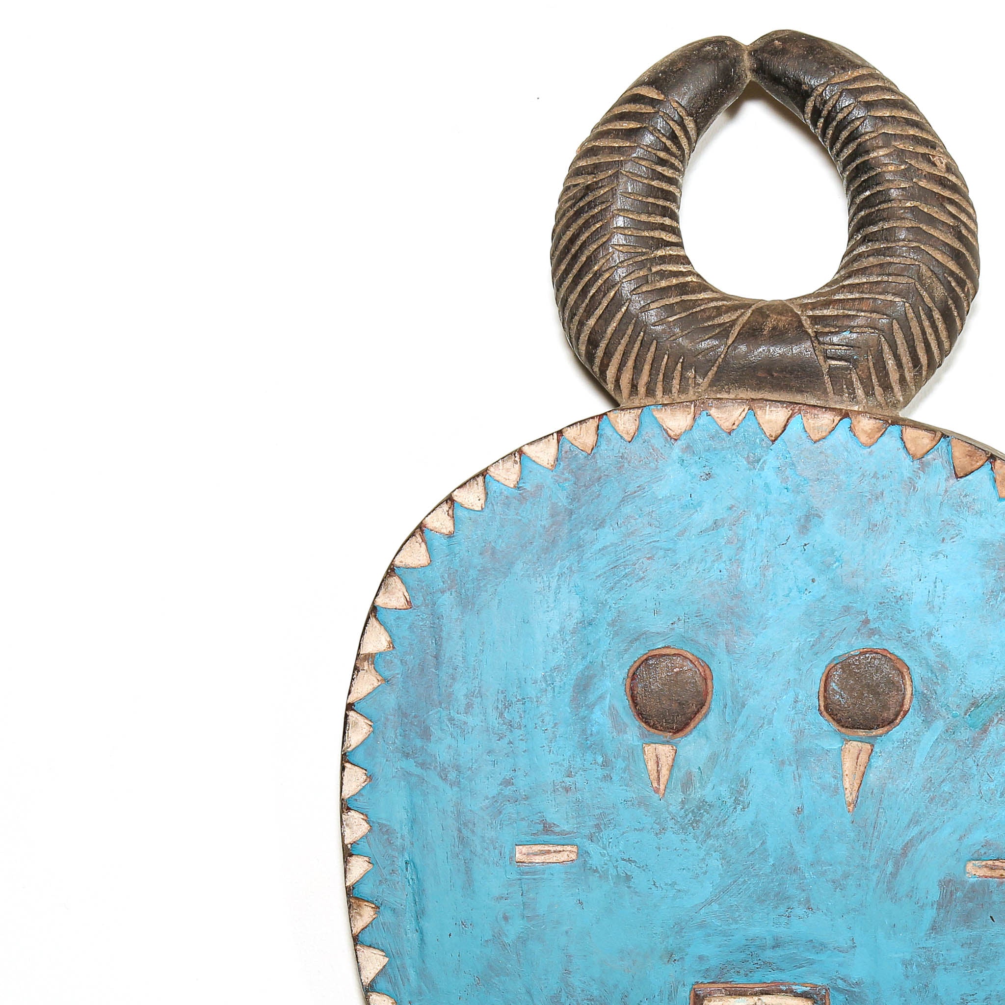 A close-up of a decorative mask or shield-like object with a bright turquoise blue face, carved dark brown horns, and small metallic gold accents on a plain white background.