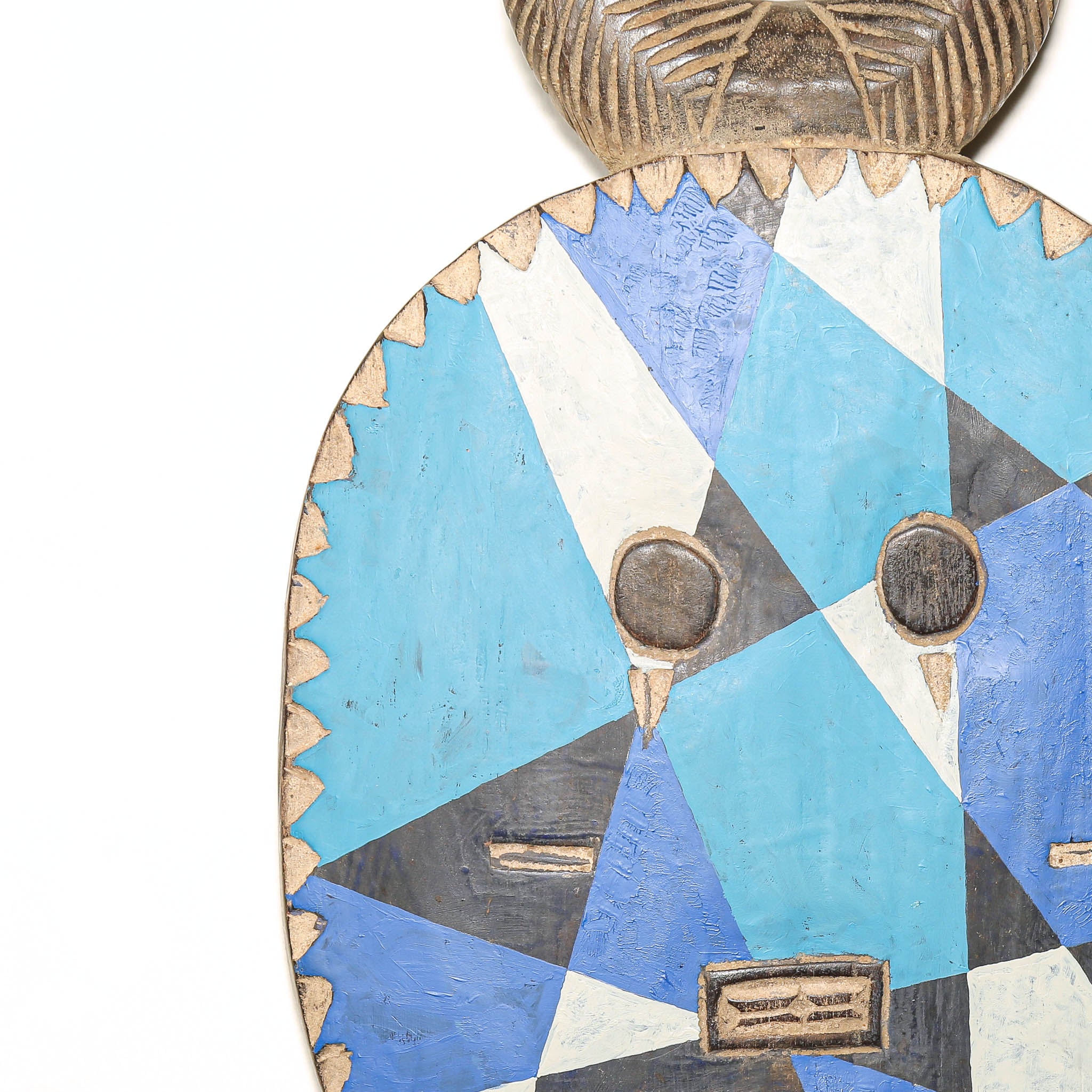 A close-up of a carved wooden mask or decorative plaque featuring a geometric face design in light blue, medium blue, white, and dark gray, with a carved textured top section visible at the upper edge against a plain white background.