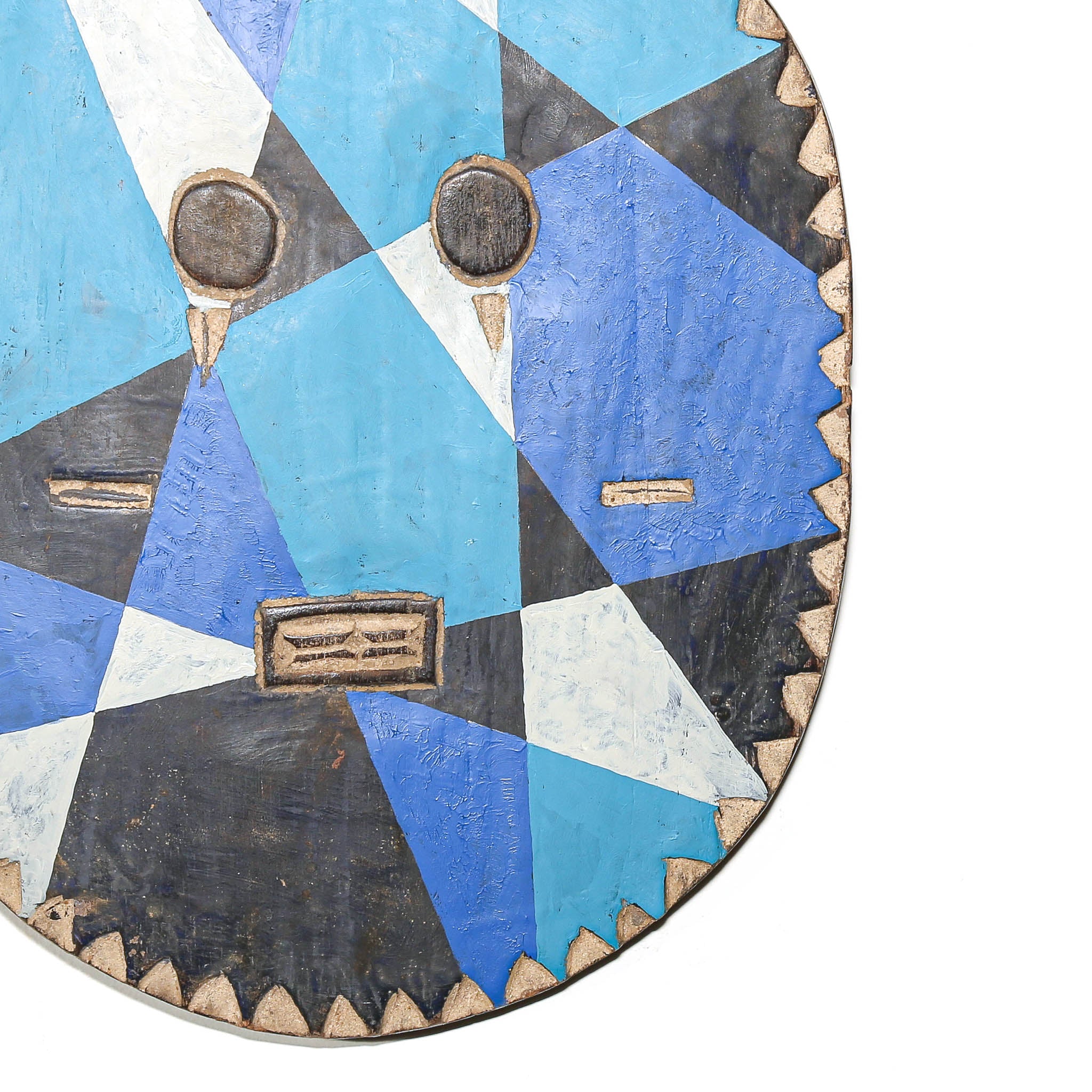 A close-up of a hand-painted wooden mask with a geometric pattern in blue, black, white, and light blue, featuring carved facial details and a serrated wooden edge.
