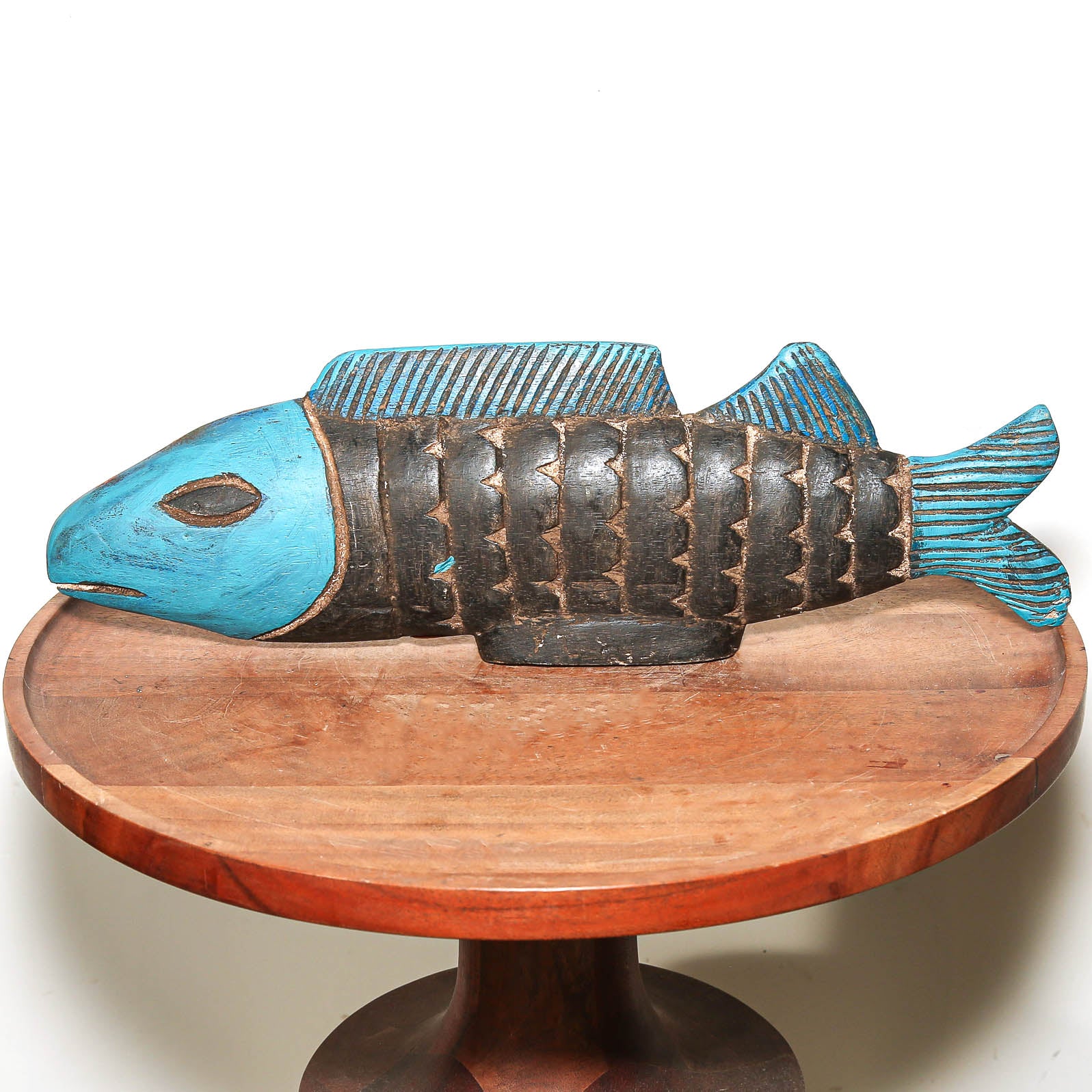 A hand-carved wooden fish sculpture with a bright turquoise blue painted head and fins, and a dark brown natural wood body, displayed on a round wooden pedestal against a white background.