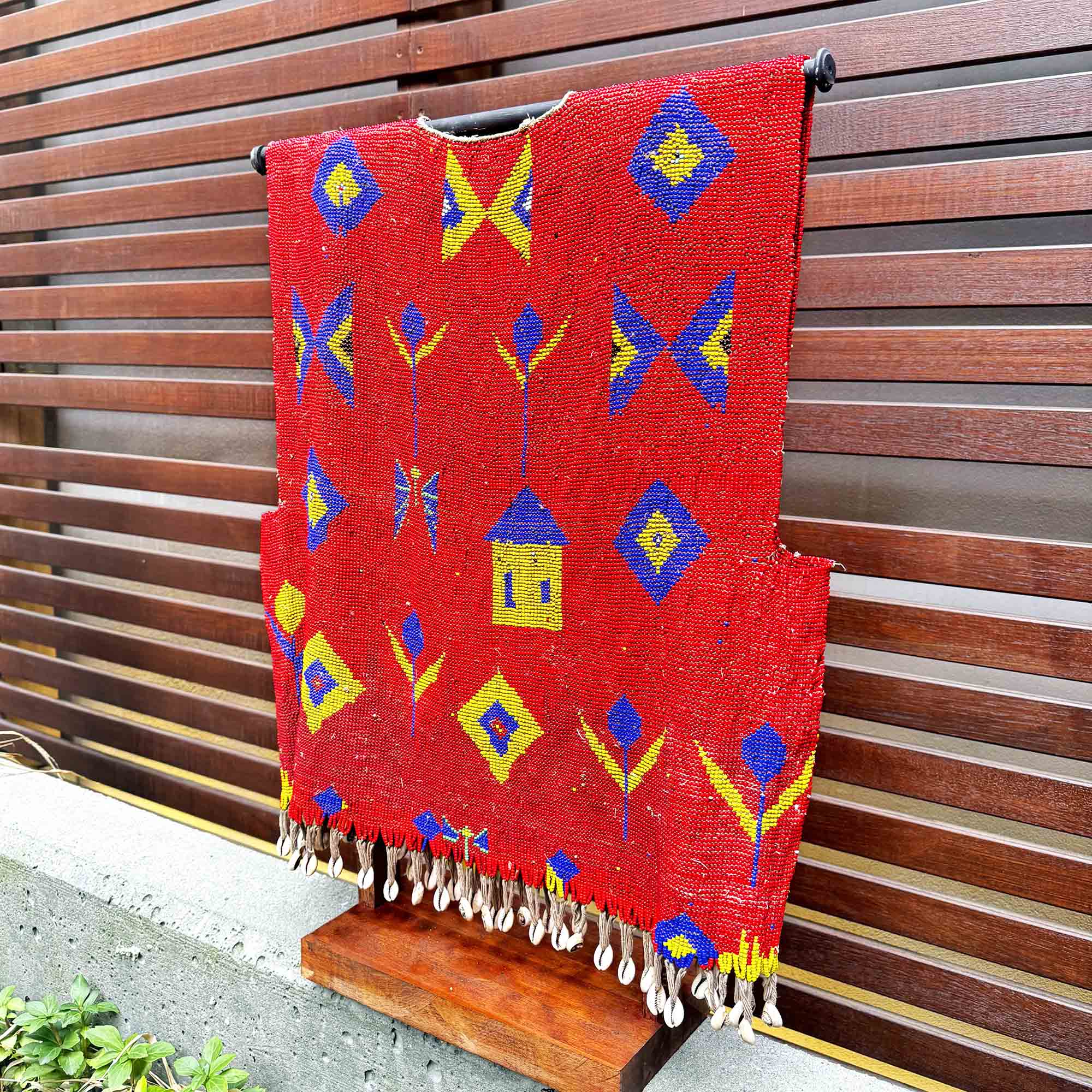 Red woven blanket with colorful patterns hanging on a wooden stand against a wooden fence.