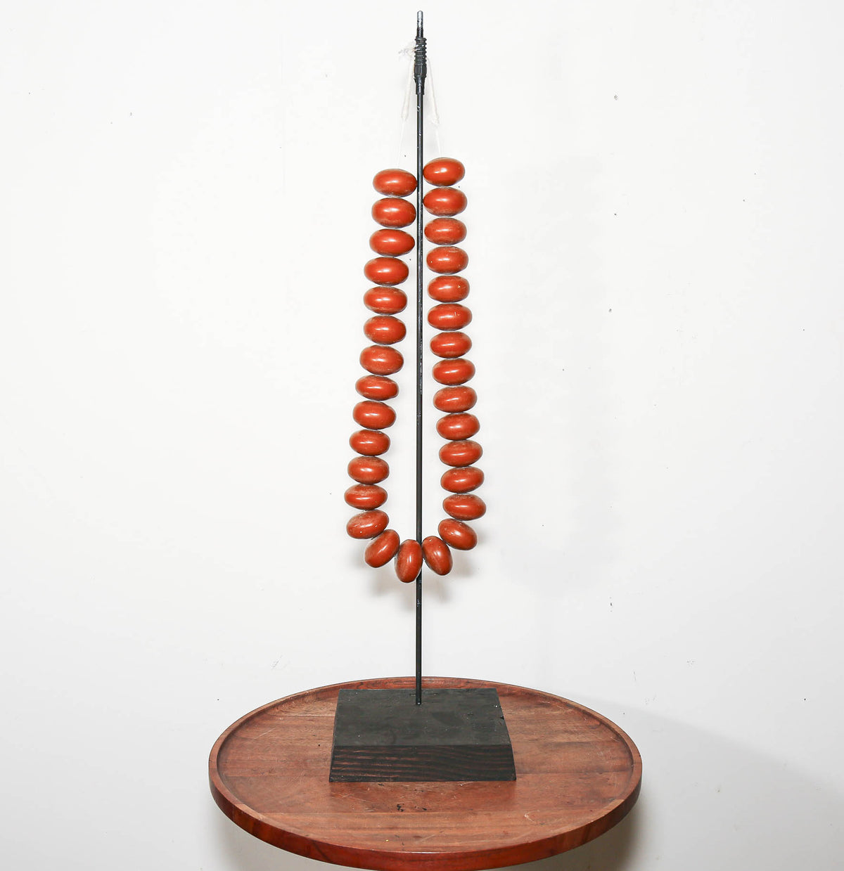 A decorative sculpture or art piece featuring two vertical strands of glossy terracotta-colored oval beads hanging from a thin metal rod, mounted on a square black base over a round wooden platform against a plain white wall.