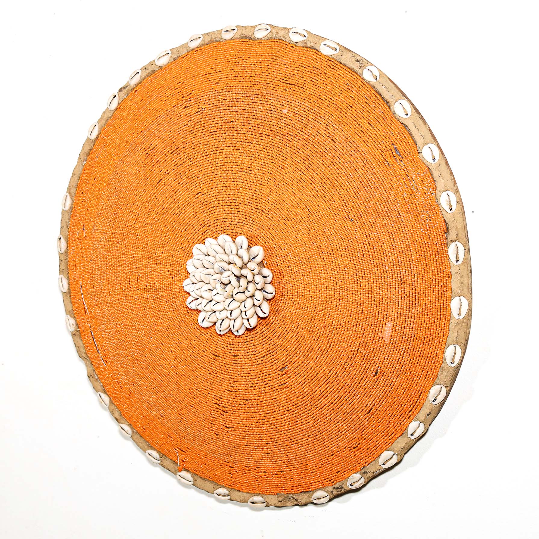 Round orange beaded wall shield decor with a central white flower design made from cowrie shells hanging on a white wall.
