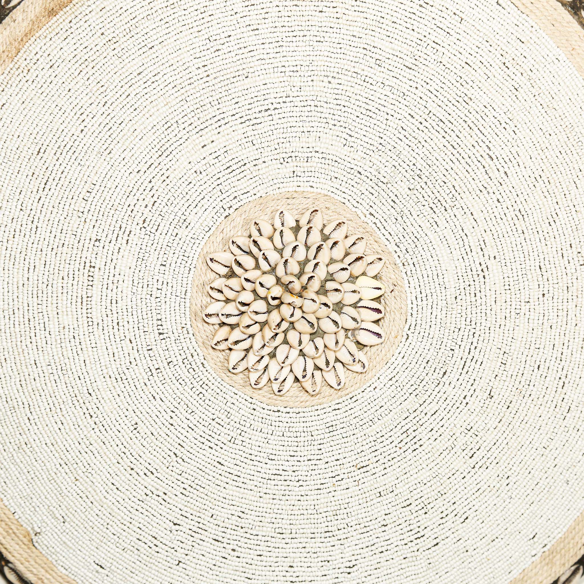 Round white beaded wall shield decor with a central white flower design made from cowrie shells hanging on a white wall.