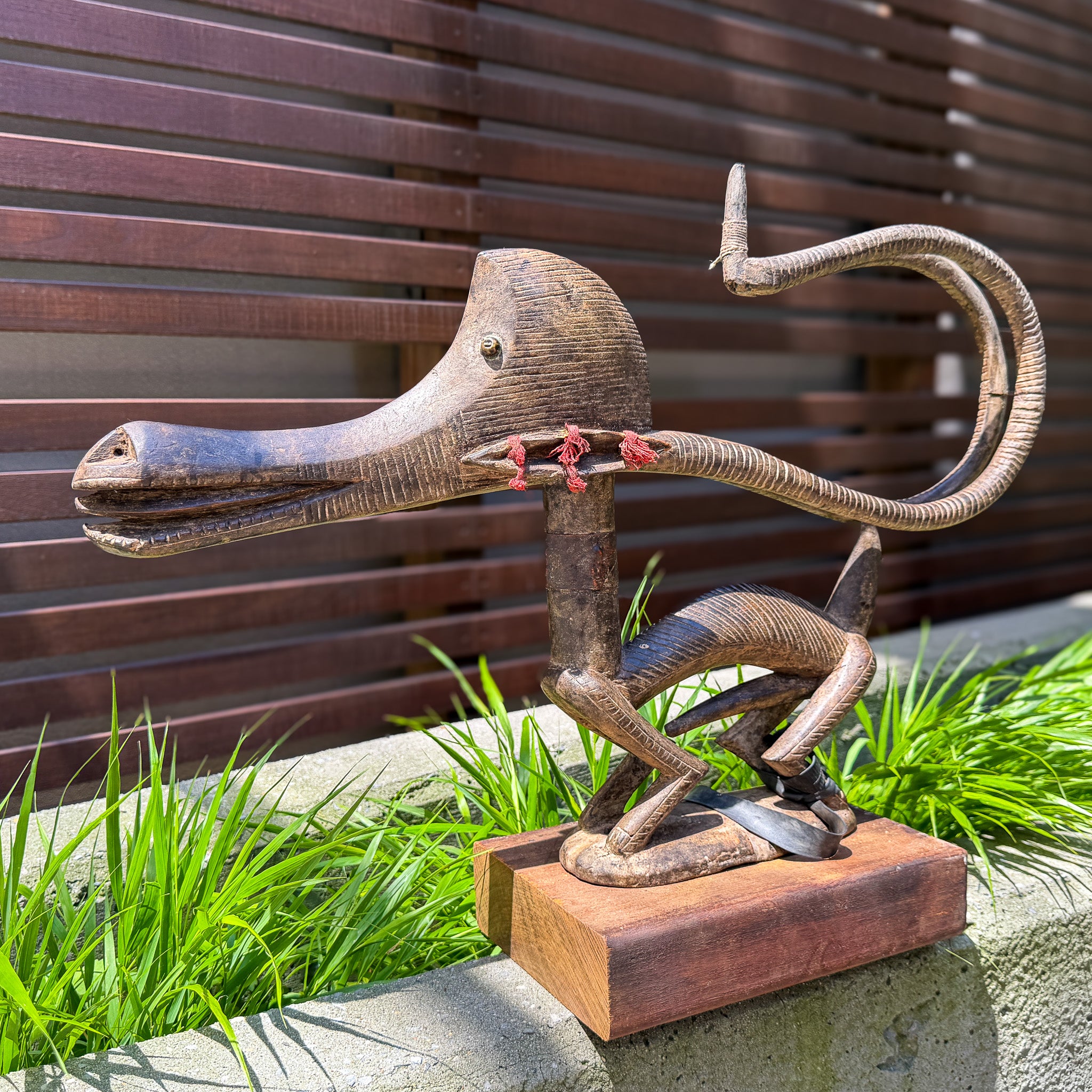 Chiwara headdress with sweeping curved horns and ridged carving. A hand-carved wooden sculpture of a stylized bird or dragon figure with a long curved beak, textured body, and red cord accents, displayed outdoors on a stone ledge with green grass and a dark brown wooden slat wall behind it.