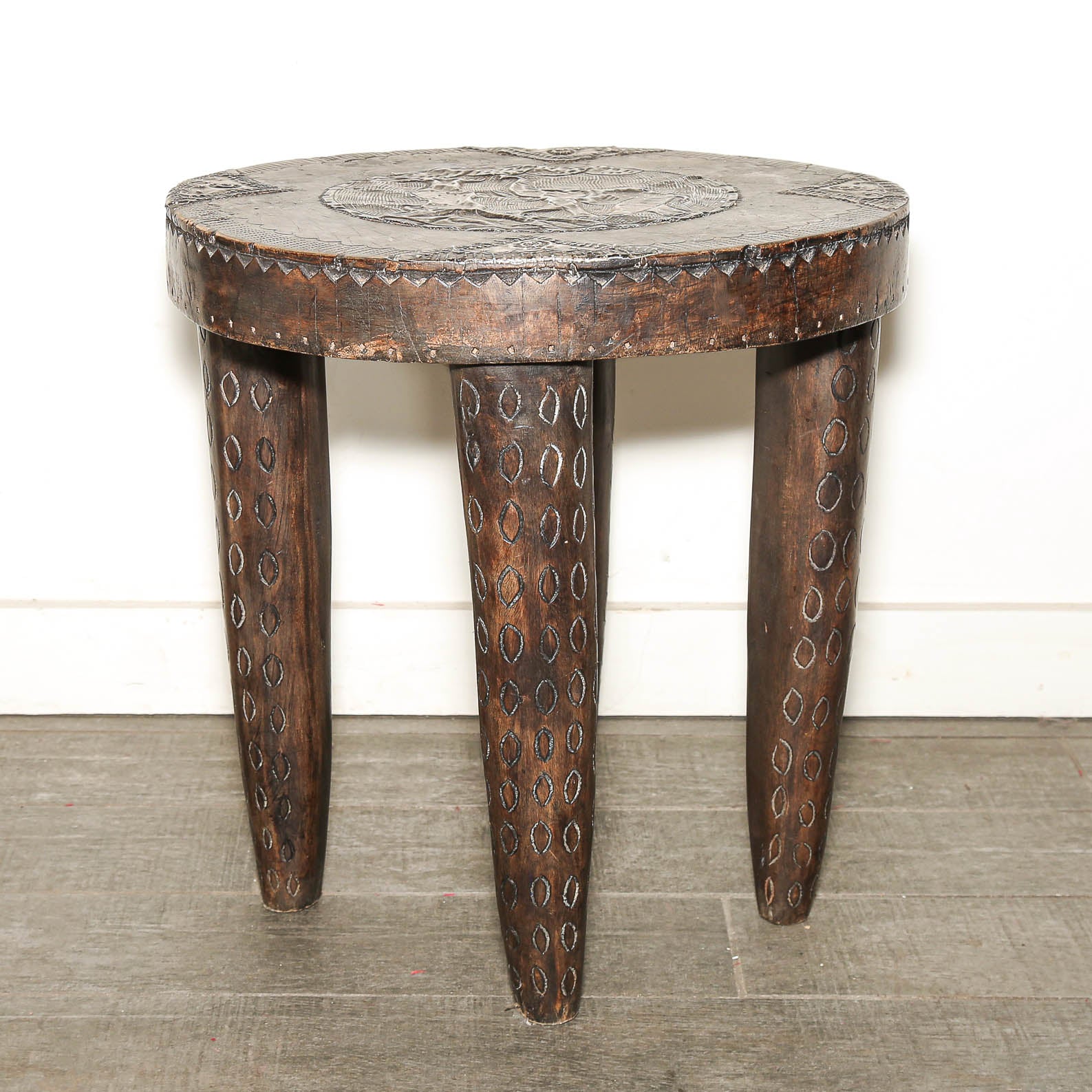 A rustic dark brown carved wooden stool with three legs, featuring circular cutout patterns on the legs and a decorative relief on the round seat, set against a plain white wall and gray floor.