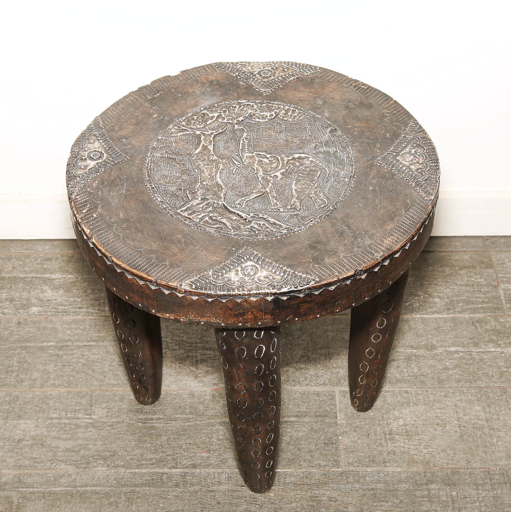 A round dark brown wooden stool with a carved animal motif on the seat and decorative cutout details on the legs, set against a white wall and gray tiled floor.