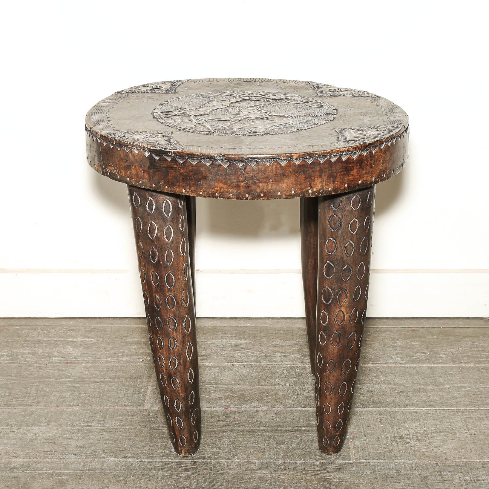 A rustic dark brown carved wooden stool with a circular seat featuring an embossed design, a beaded rim, and three tapered legs decorated with carved geometric patterns, set against a white wall and gray floor.