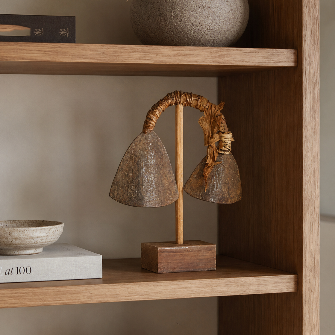 A rustic African sculpture metallic bell-shaped shades on a wooden stand, displayed on a wooden bookshelf alongside books and ceramic decor.