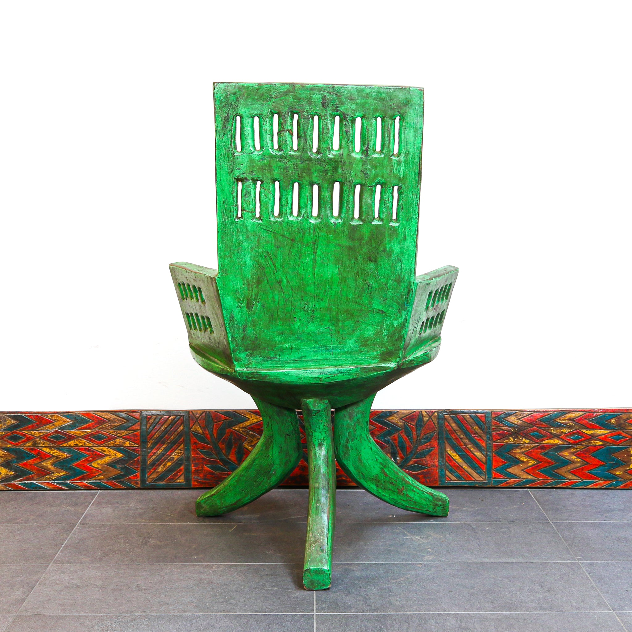 A green wooden armchair with a distressed finish, featuring a slatted backrest and curved legs, set against a white wall with a patterned tile baseboard.