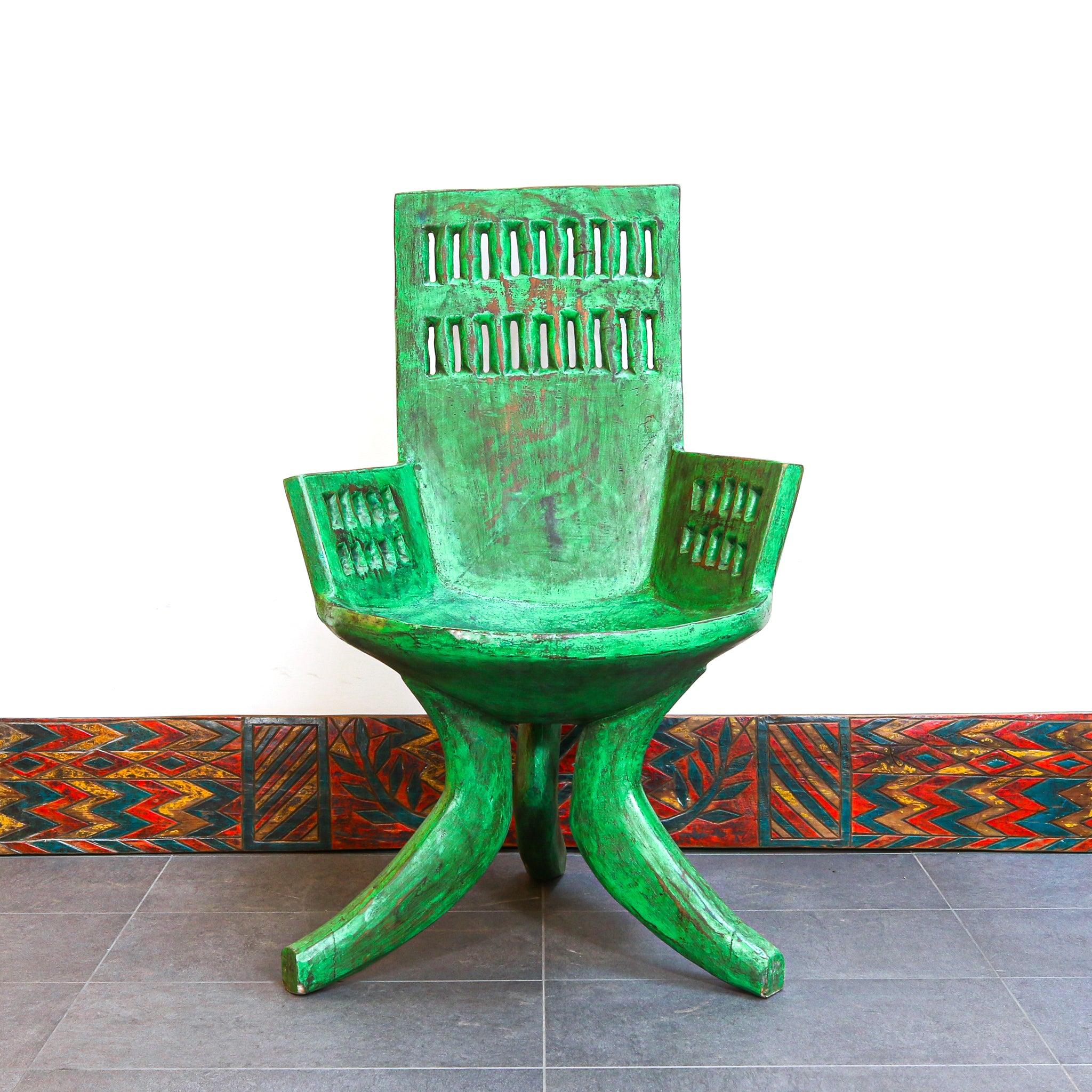 A green wooden chair with a distressed, weathered finish, featuring a high slatted backrest, armrests, and three curved legs, set against a white wall and a patterned tile baseboard.
