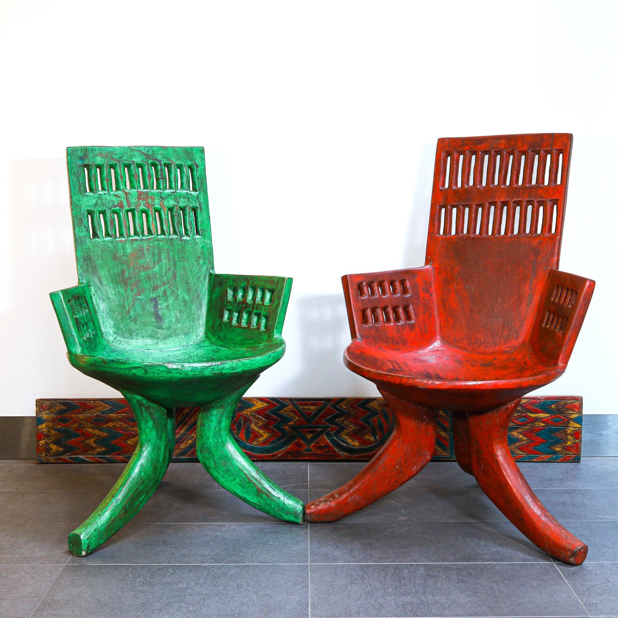 Two sculptural wooden lounge chairs with distressed painted finishes, one in green and one in red, displayed side-by-side against a white wall.