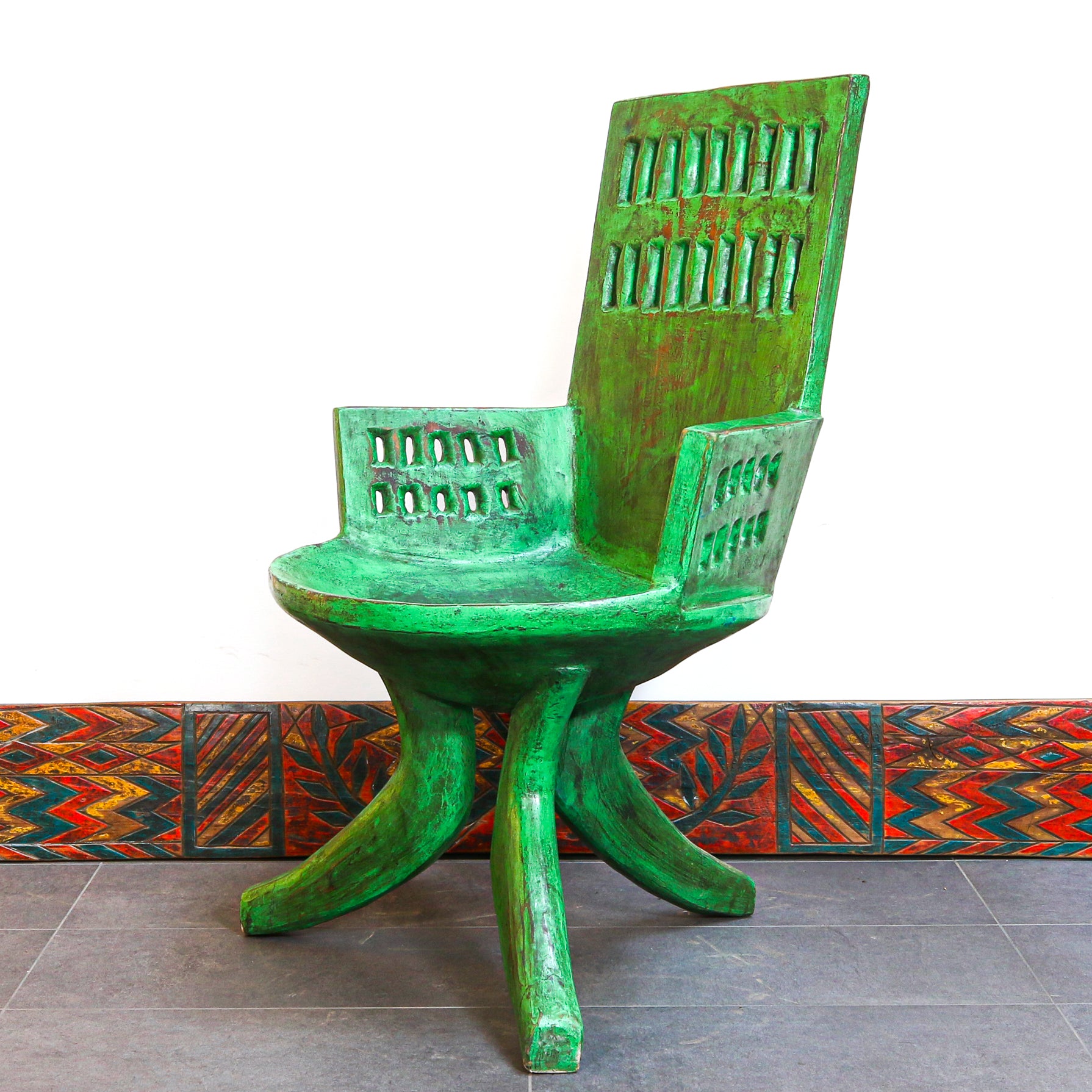 A rustic green wooden chair with a distressed finish, featuring a high slatted backrest and armrests, set against a white wall with a colorful patterned tile base.