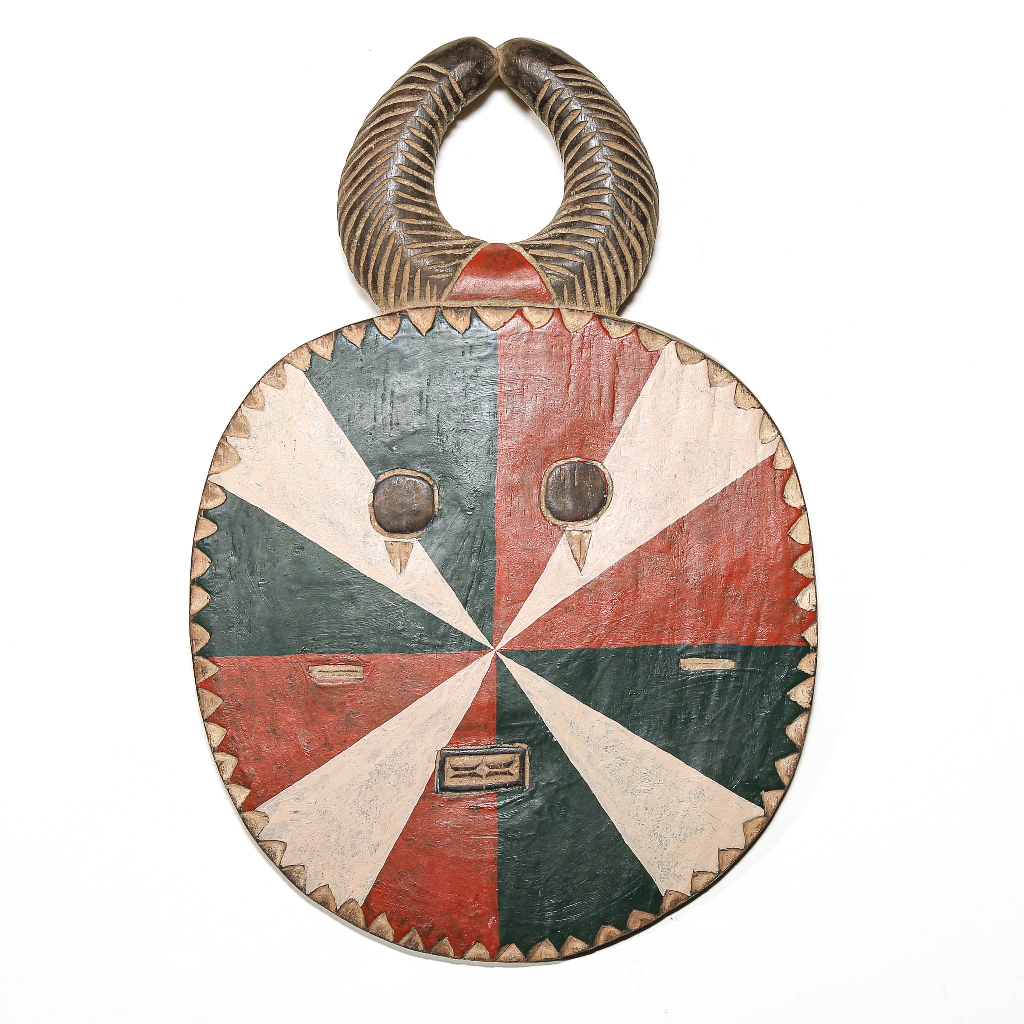 A hand-carved African wooden mask with a circular face, painted in alternating red, cream, and dark green segments, featuring a curved horn-like handle at the top.