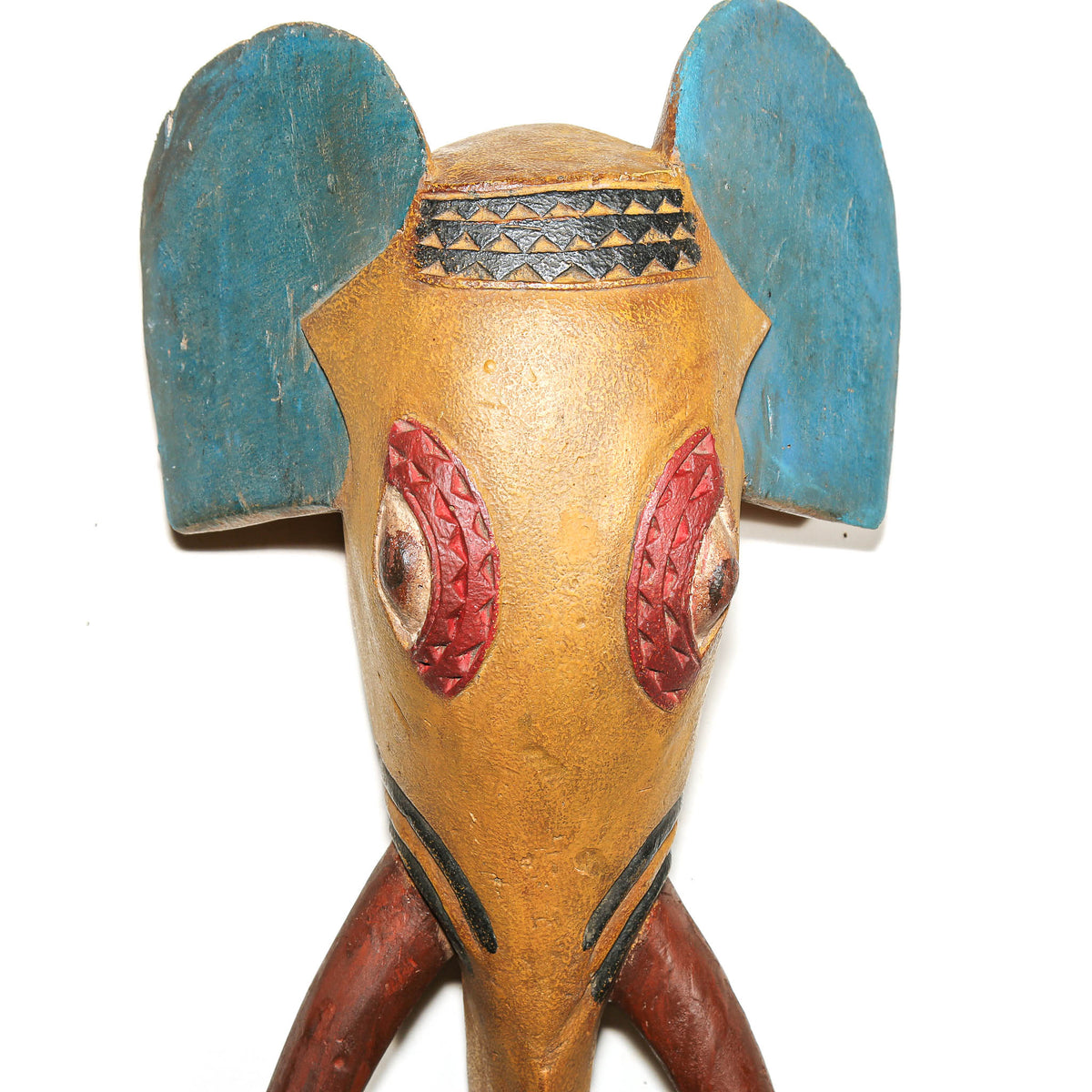 A handcrafted wooden elephant head figurine with a weathered tan-brown body, large blue ears, red patterned eyes, and black decorative details, shown against a plain white background.