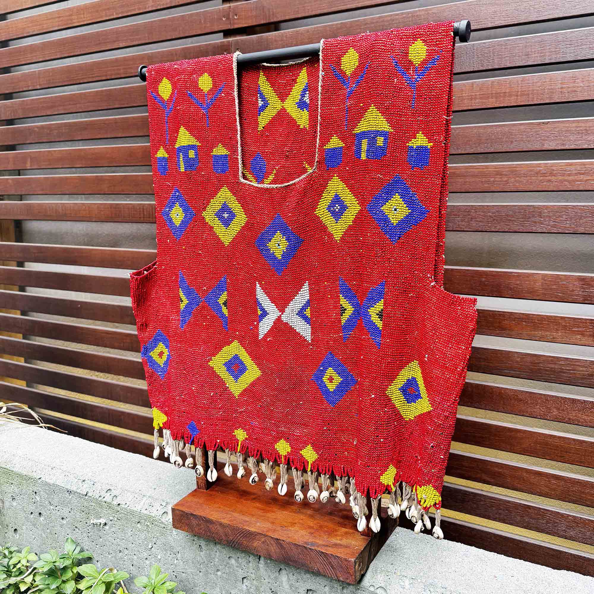 Red african beads shirt with colorful geometric patterns on a wooden stand against a wooden slat background