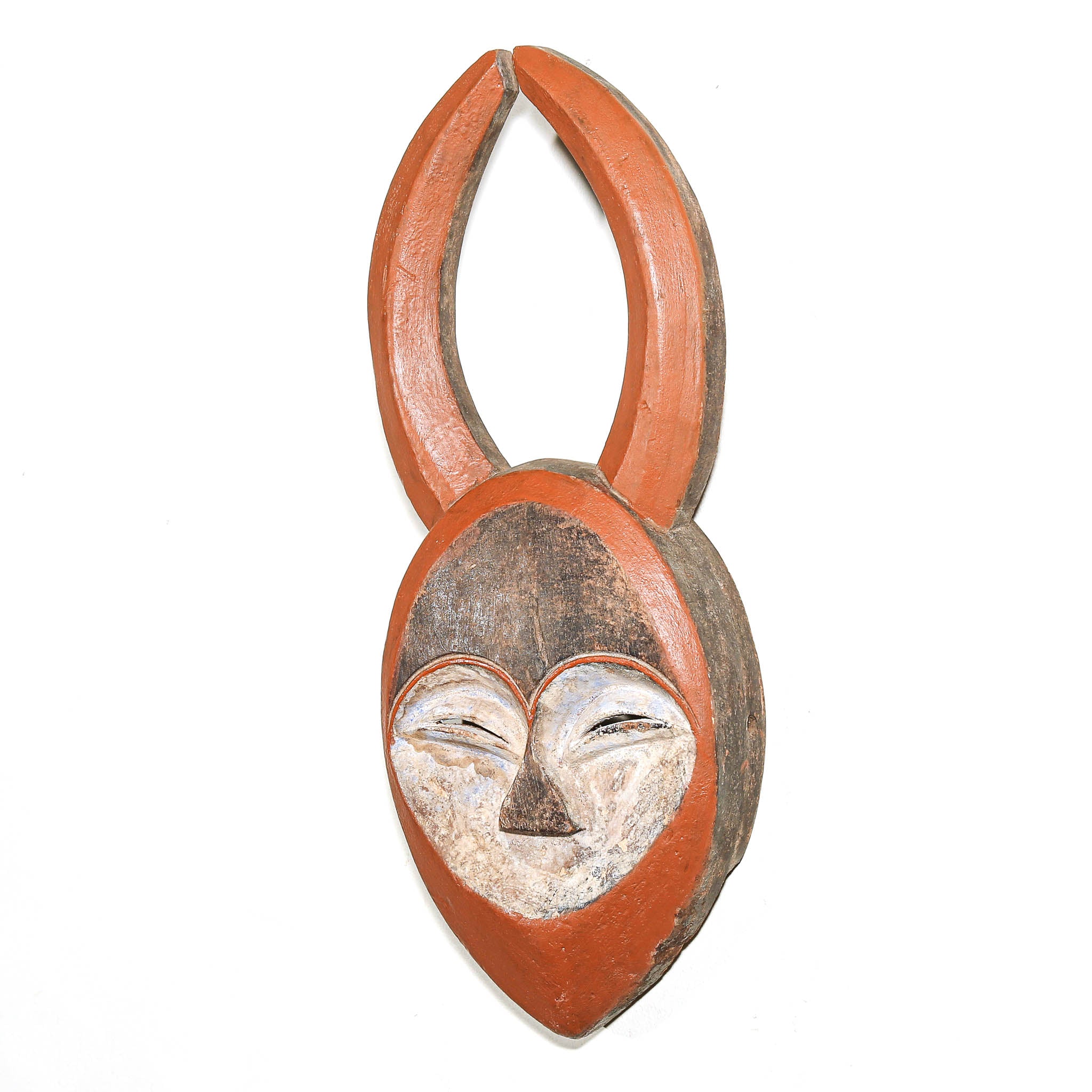 A handcrafted wooden wall hanging sculpture shaped like a stylized face with curved antler-like extensions at the top, painted in terracotta orange, brown, cream, and black on a plain white background.