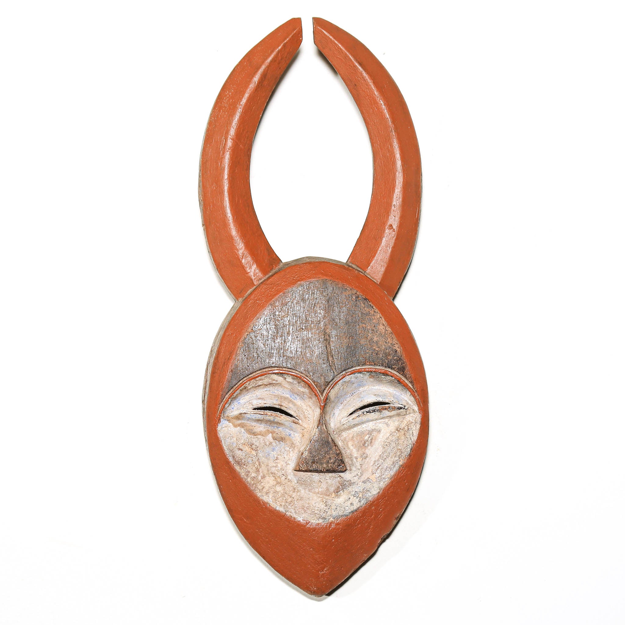 A hand-carved wooden mask with a reddish-brown painted finish, featuring a stylized face with closed eyes and curved horn-like extensions at the top, shown against a plain white background.