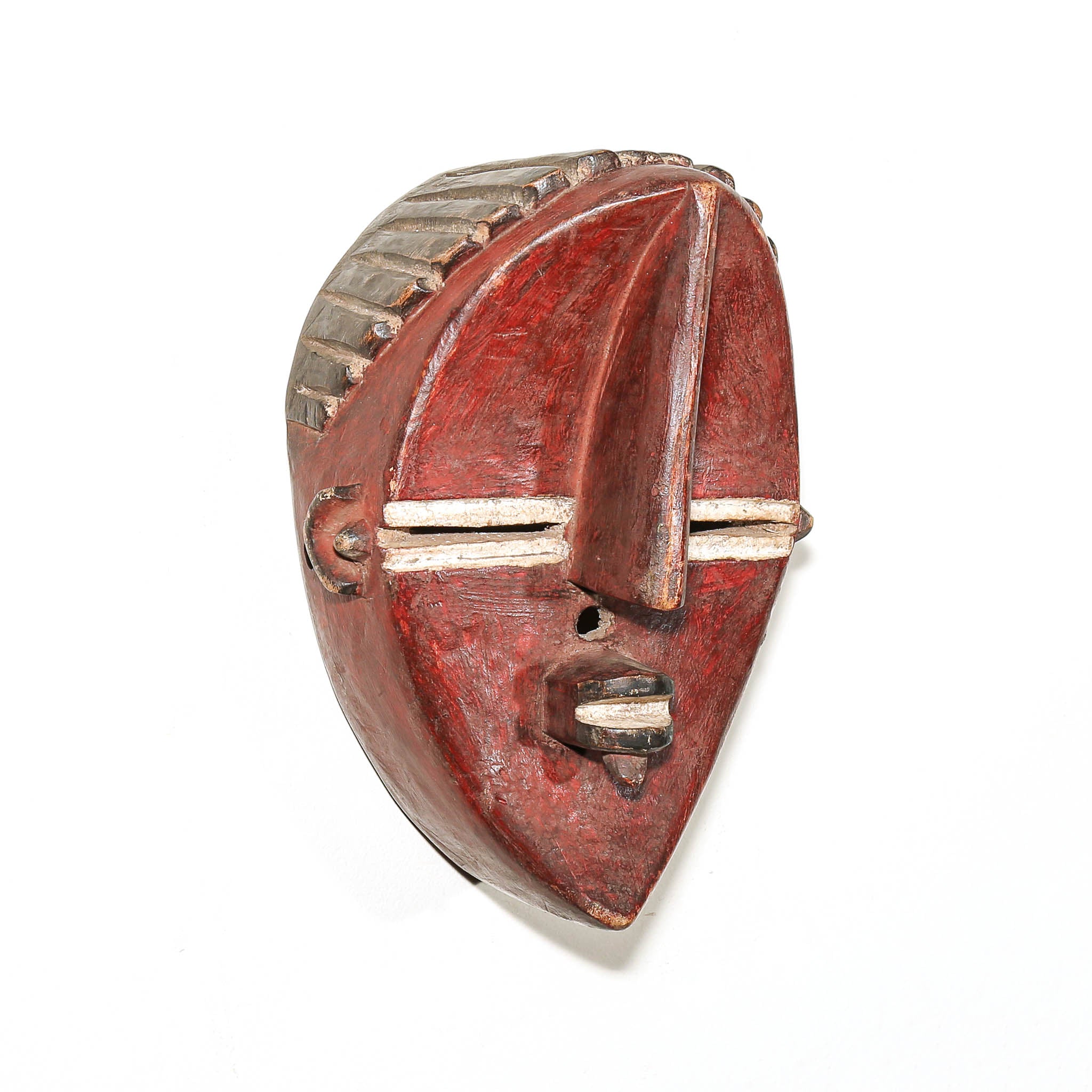 A carved wooden mask with a reddish-brown body, featuring a prominent nose, horizontal eye slits with white accents, and a textured crown, photographed on a white background.