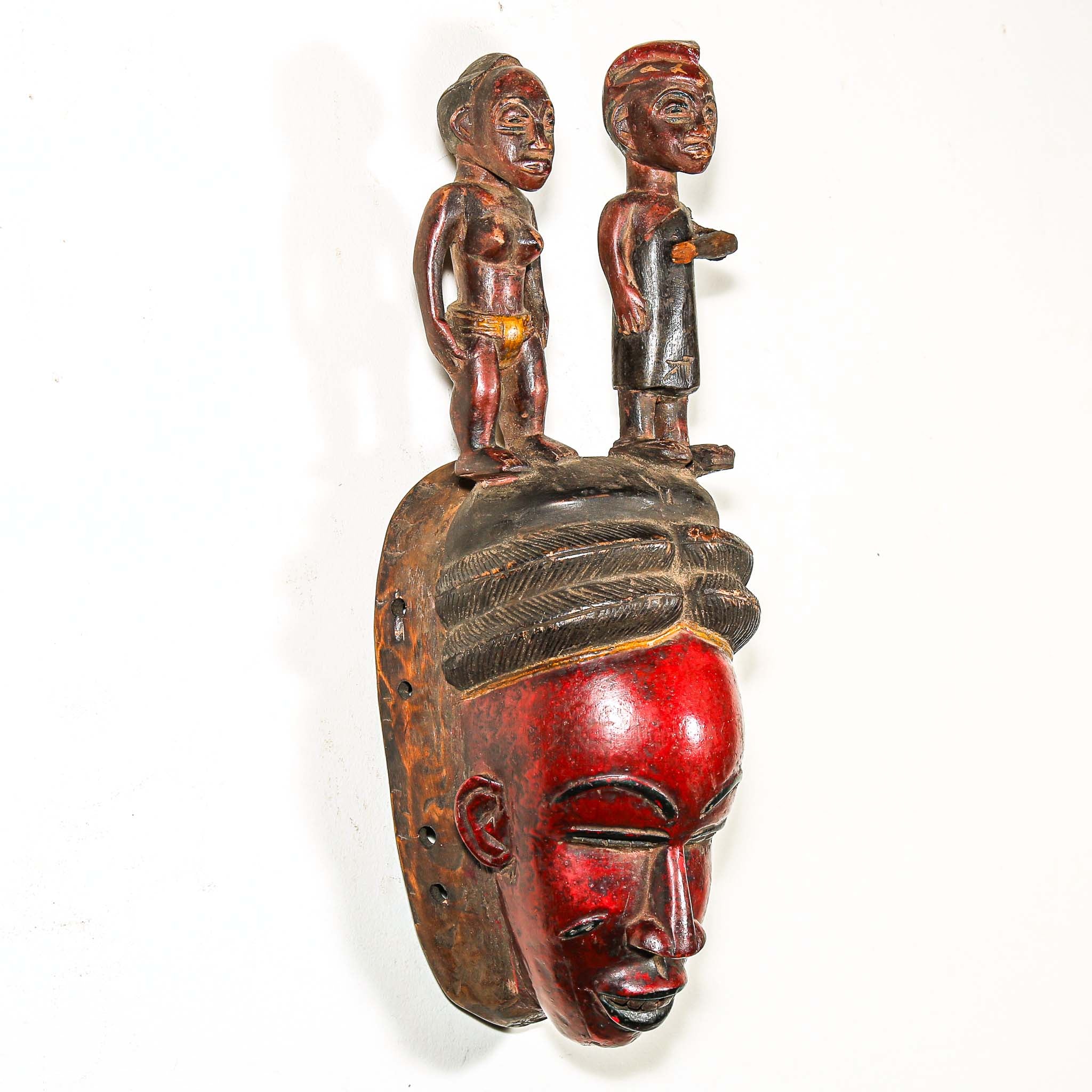 Wooden mask with a red face and two figures on top against a white background