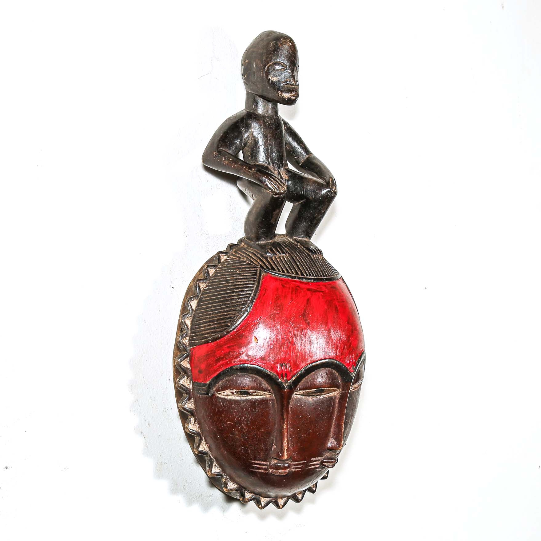 African mask with a red face and a figure on top against a white background