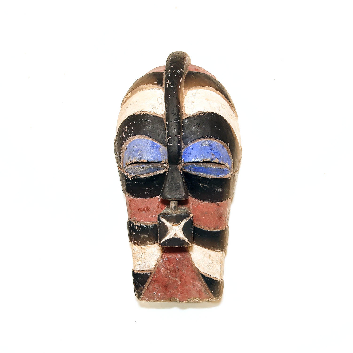 A small carved decorative mask with a black handle on top, featuring geometric patterns in black, white, reddish-brown, and blue on a plain white background.