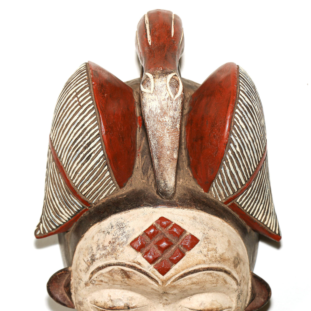 A carved wooden African-style mask or sculpture with a bird on its head, featuring red-painted sections, striped beige and brown wings, and a red diamond pattern on the forehead, shown against a plain white background.