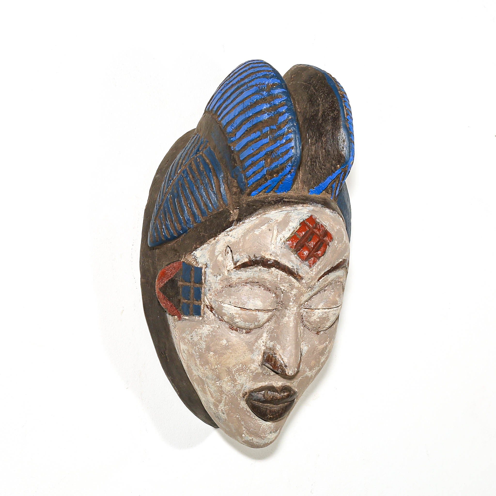 A carved wooden African-style mask with a stylized face, blue striped hair, and red geometric accents, photographed against a plain white background.