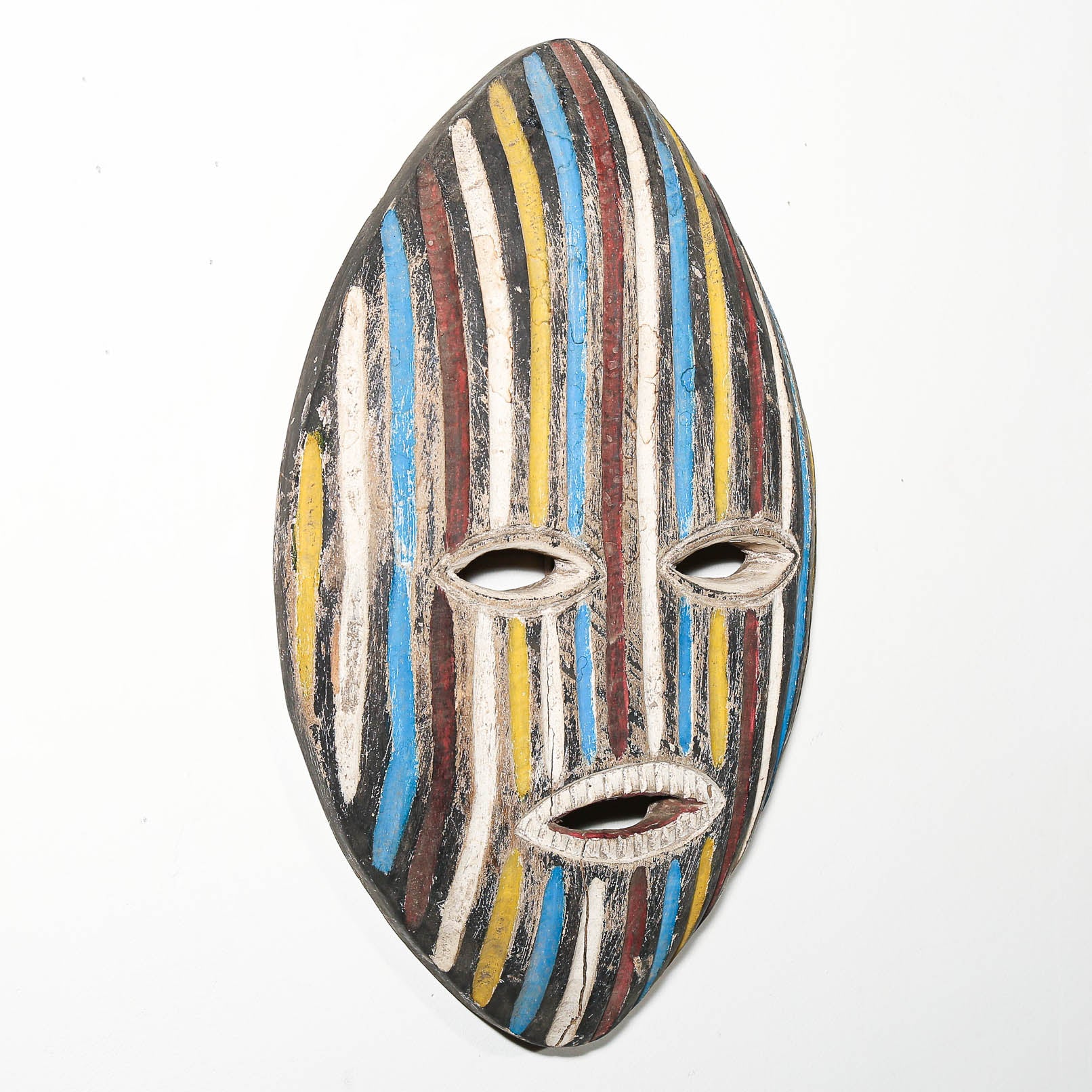 A handcrafted wooden mask with a vertically striped pattern in blue, yellow, brown, white, and black, featuring cutouts for the eyes and mouth, shown against a plain white background.