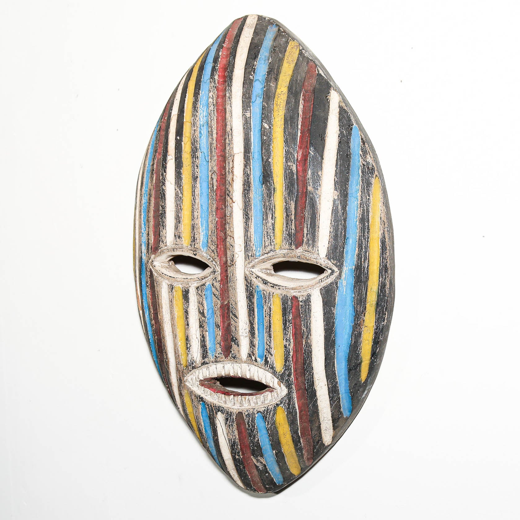 A hand-carved wooden mask with a vertically elongated shape, featuring painted vertical stripes in blue, yellow, red, white, and black, set against a plain white background.