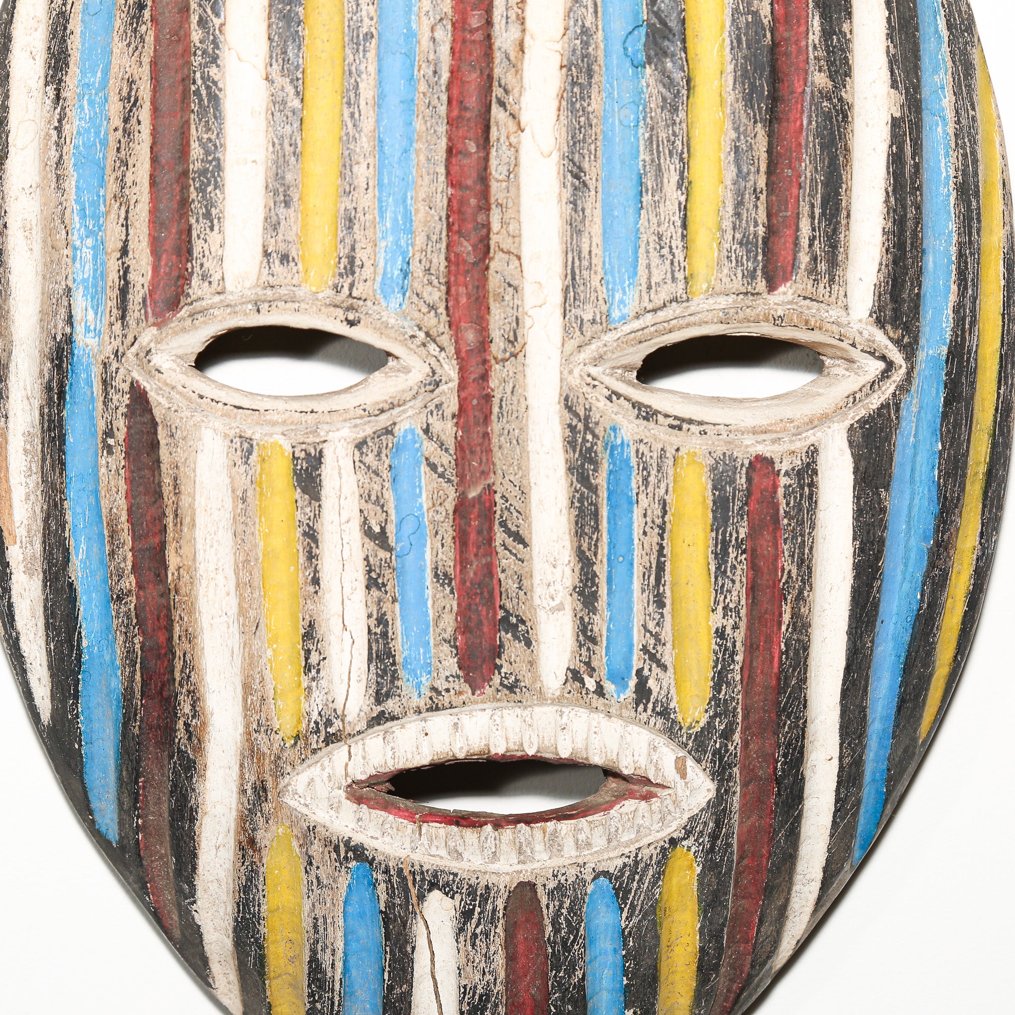 A close-up of a hand-painted wooden mask with vertical stripes in blue, yellow, red, black, and off-white, featuring cutouts for the eyes and mouth.