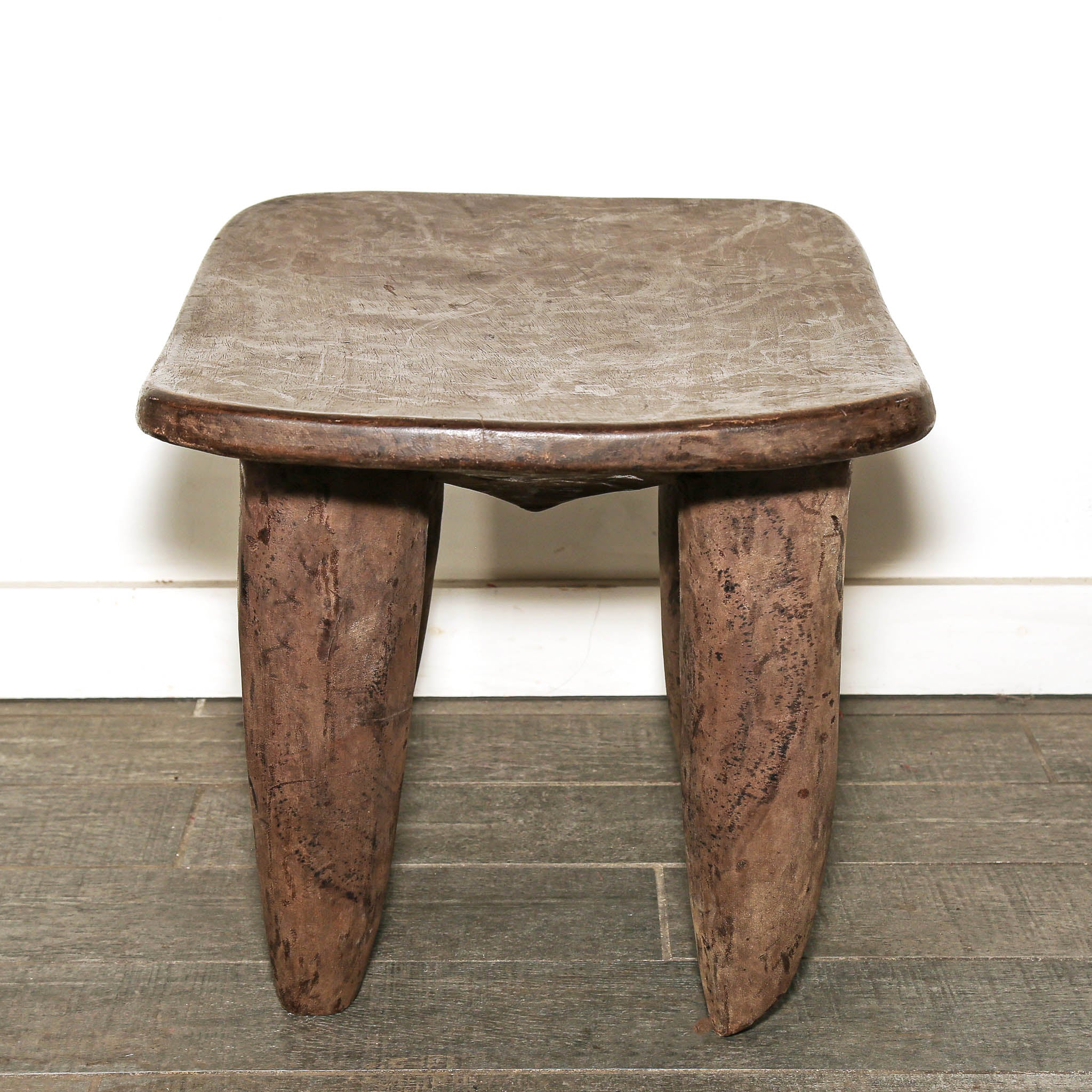 African Senufo table with sculptural curved surface and slender wooden legs. A rustic carved wooden stool with a square seat and two thick tapered legs, shown against a white wall and gray wood floor.