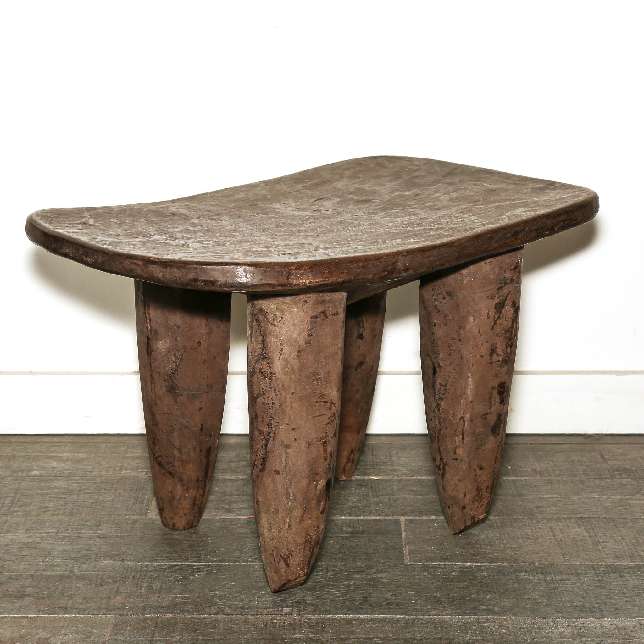 Traditional Senufo wood table from Ivory Coast with distinctive curved design. A rustic dark brown wooden stool with a curved, organic-shaped seat and three thick, tapered legs, set against a white wall and dark wood floor.