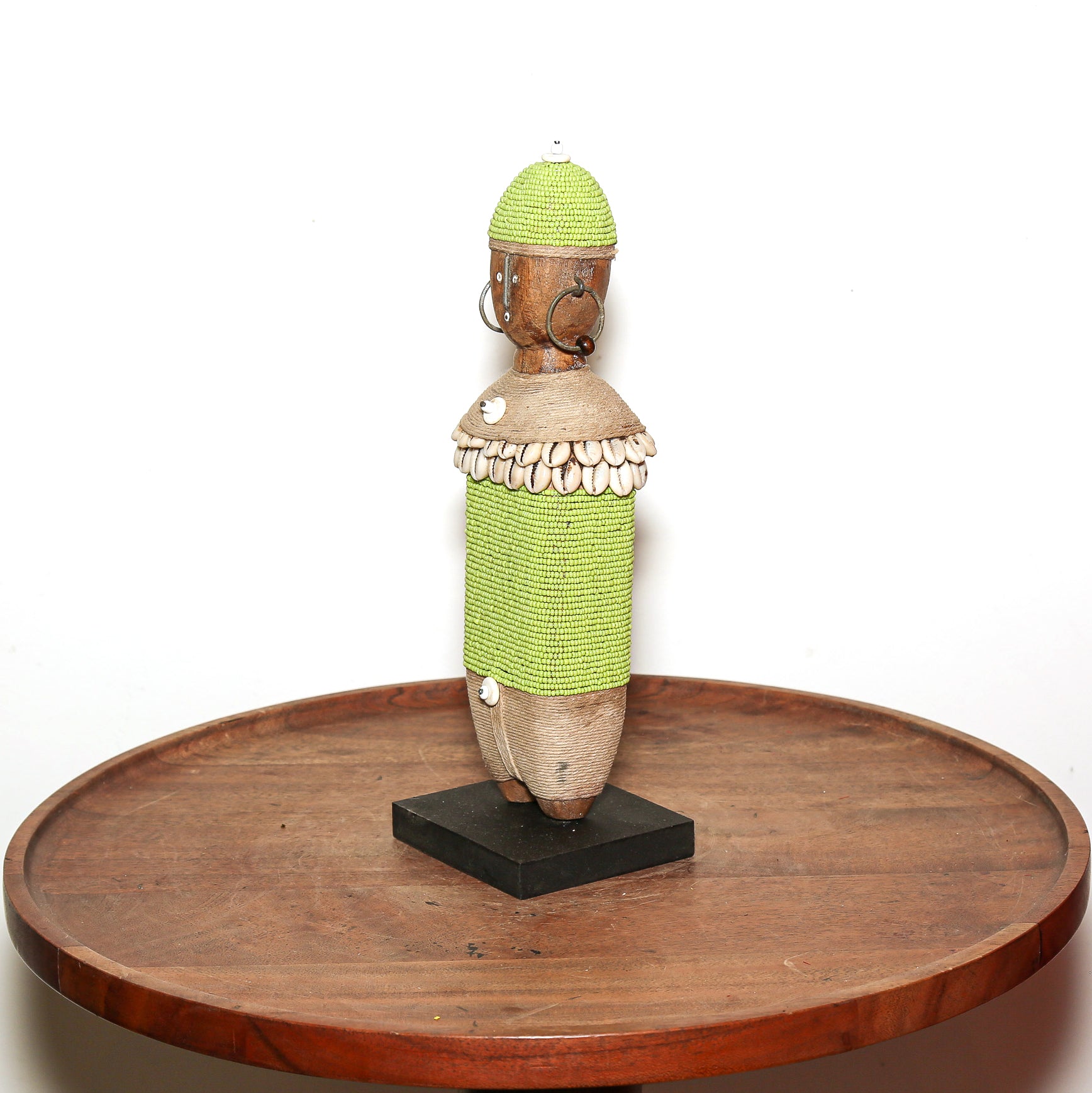 A handmade African-style wooden doll sculpture with a green beaded torso and hat, mounted on a black base and displayed on a round wooden table against a white background.