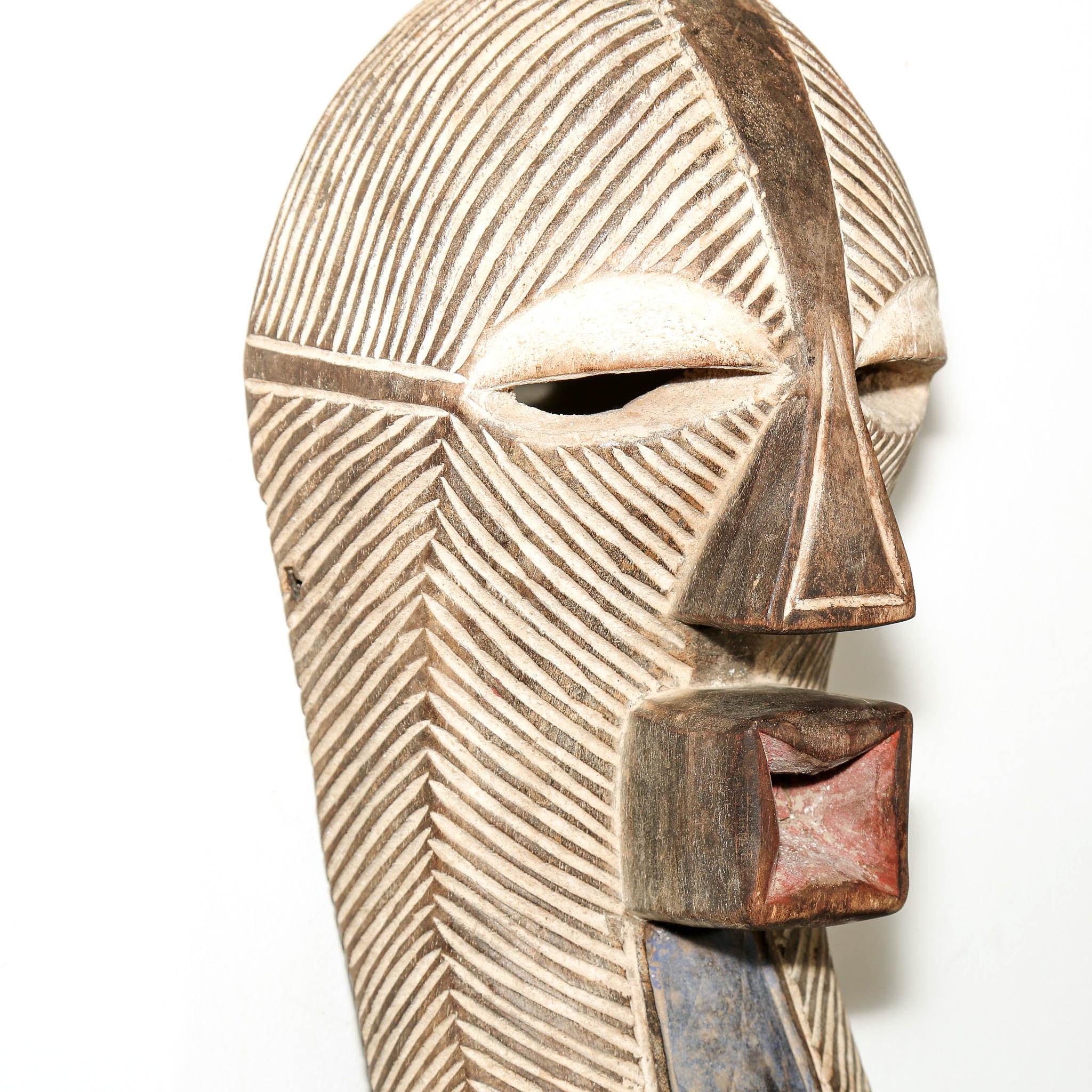 A close-up of a carved wooden mask with geometric facial features, hand-dyed details, and textured linear patterns on a plain white background.