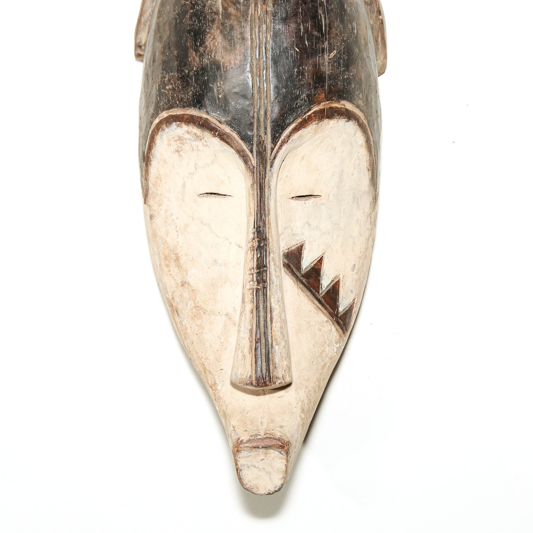 A hand-carved wooden mask with a stylized face, featuring a dark brown top section and a lighter beige lower section with carved facial details, set against a plain white background.