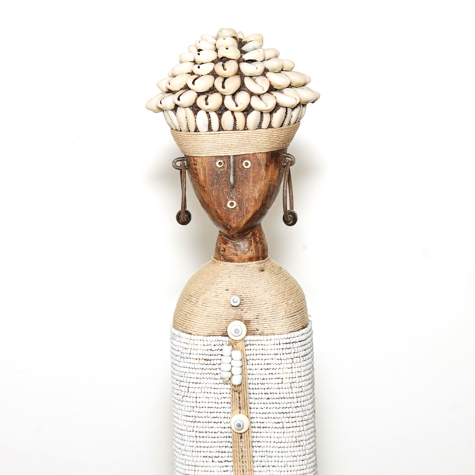 A handmade African-style wooden doll sculpture with a carved brown face, a woven beige headband decorated with white cowrie shells, dangling earrings, and a textured white and beige body, set against a plain white background.