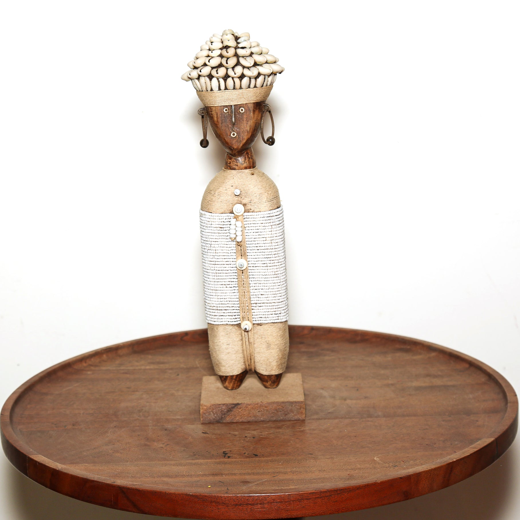 A hand-carved African wooden figurine of a woman with a beaded hat and waist, displayed on a round wooden tray against a white background.
