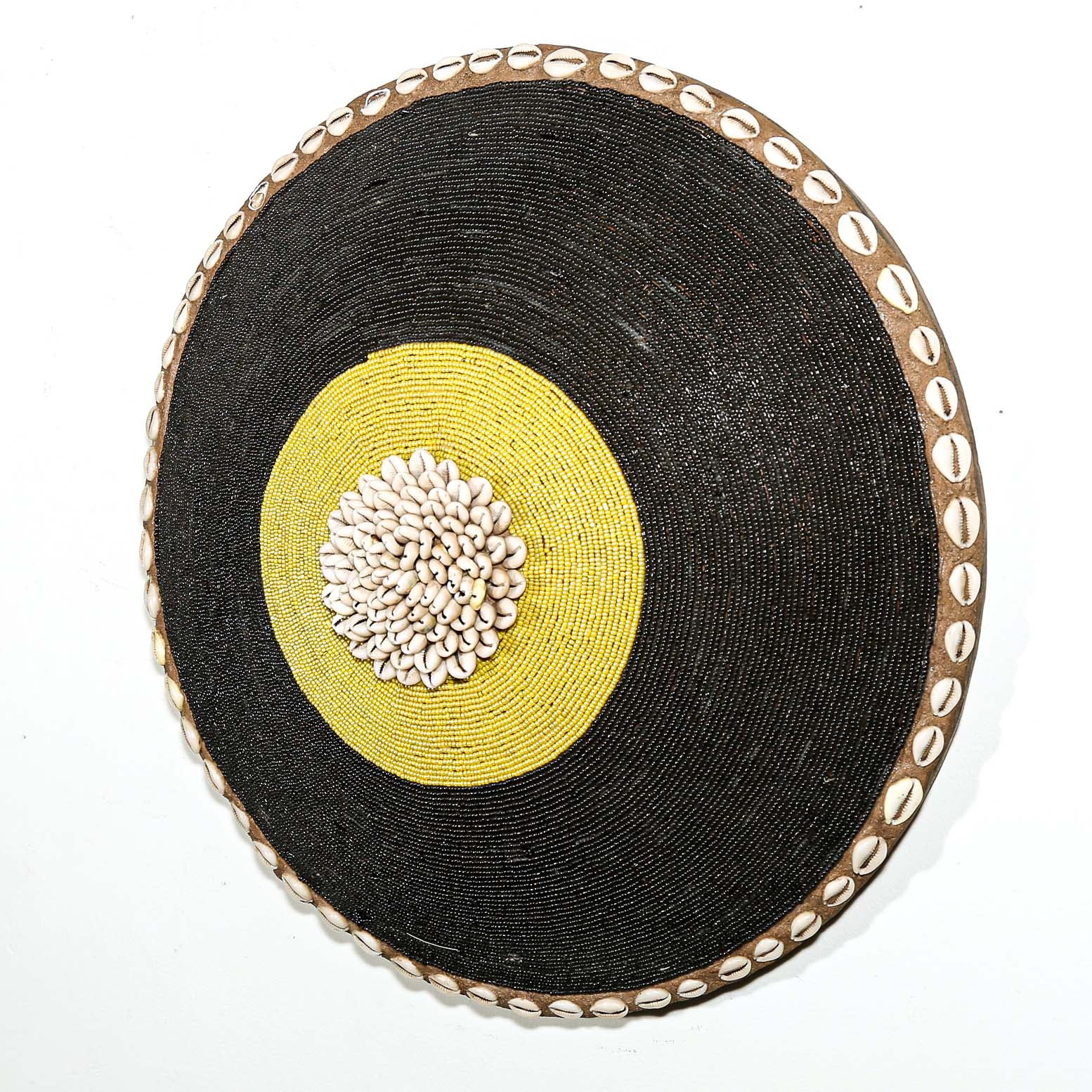Round woven beaded shield with black surface, yellow center, and decorative edge hanging on a white wall