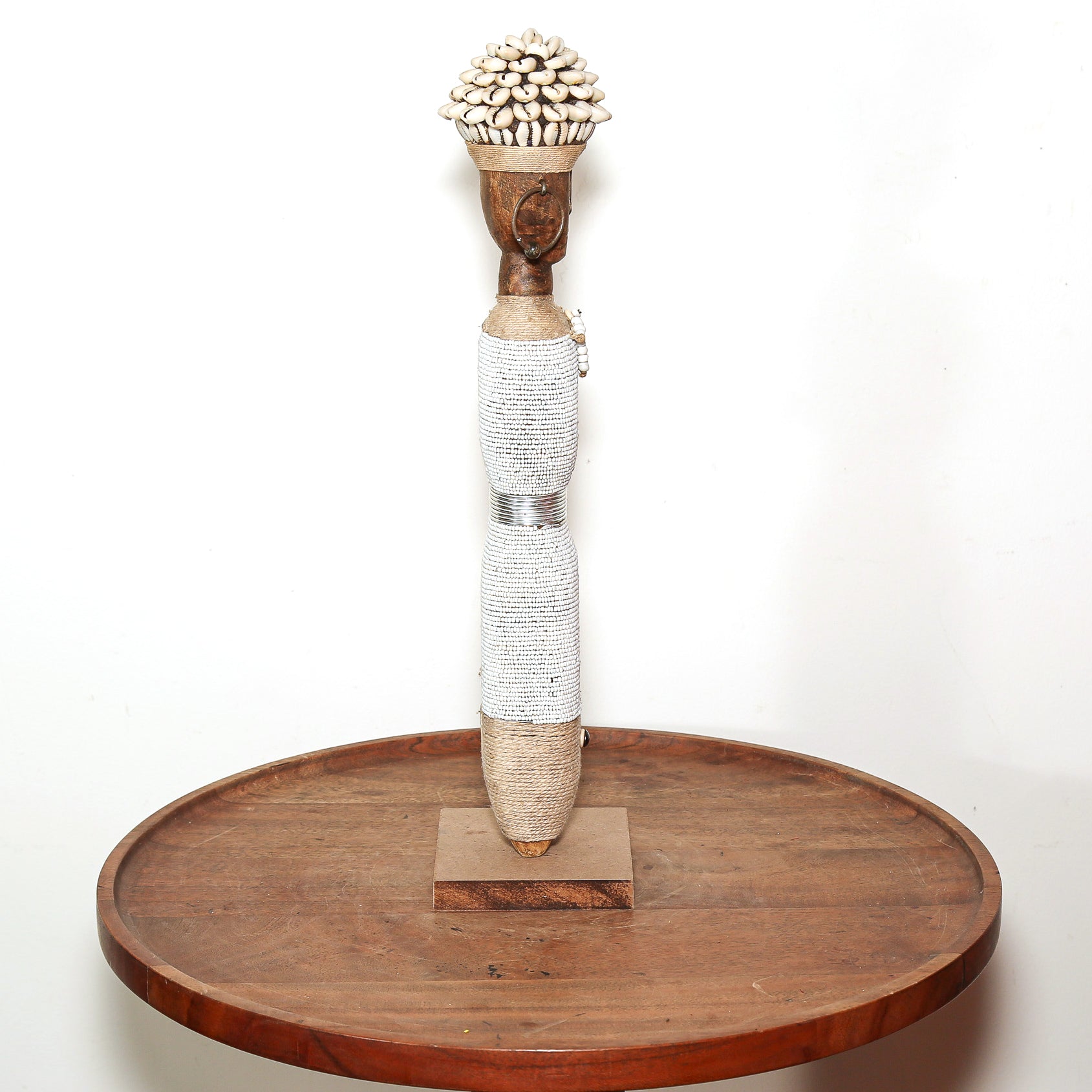 A handmade African-style wooden pole art sculpture with a woven body and a cluster of small cowrie shells on the top, displayed on a round wooden tray against a white background.
