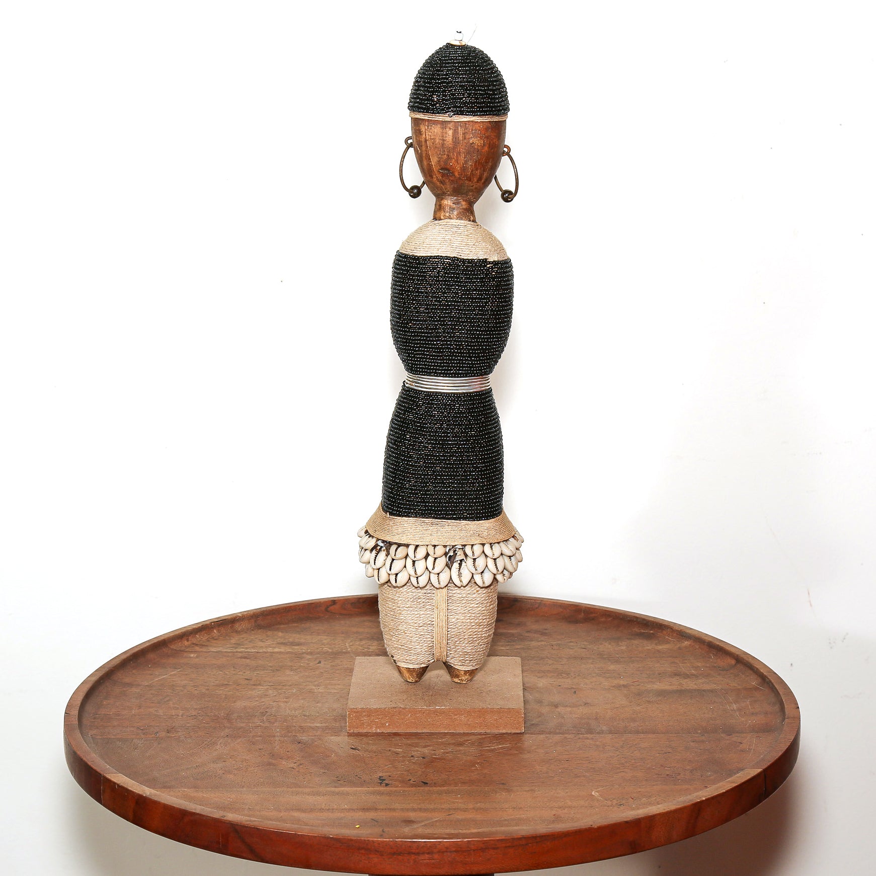 A tall, narrow African-style decorative figurine sculpture with a black beaded body, natural wood head and legs, and shell-like detailing at the waist, displayed on a round wooden pedestal against a white background.